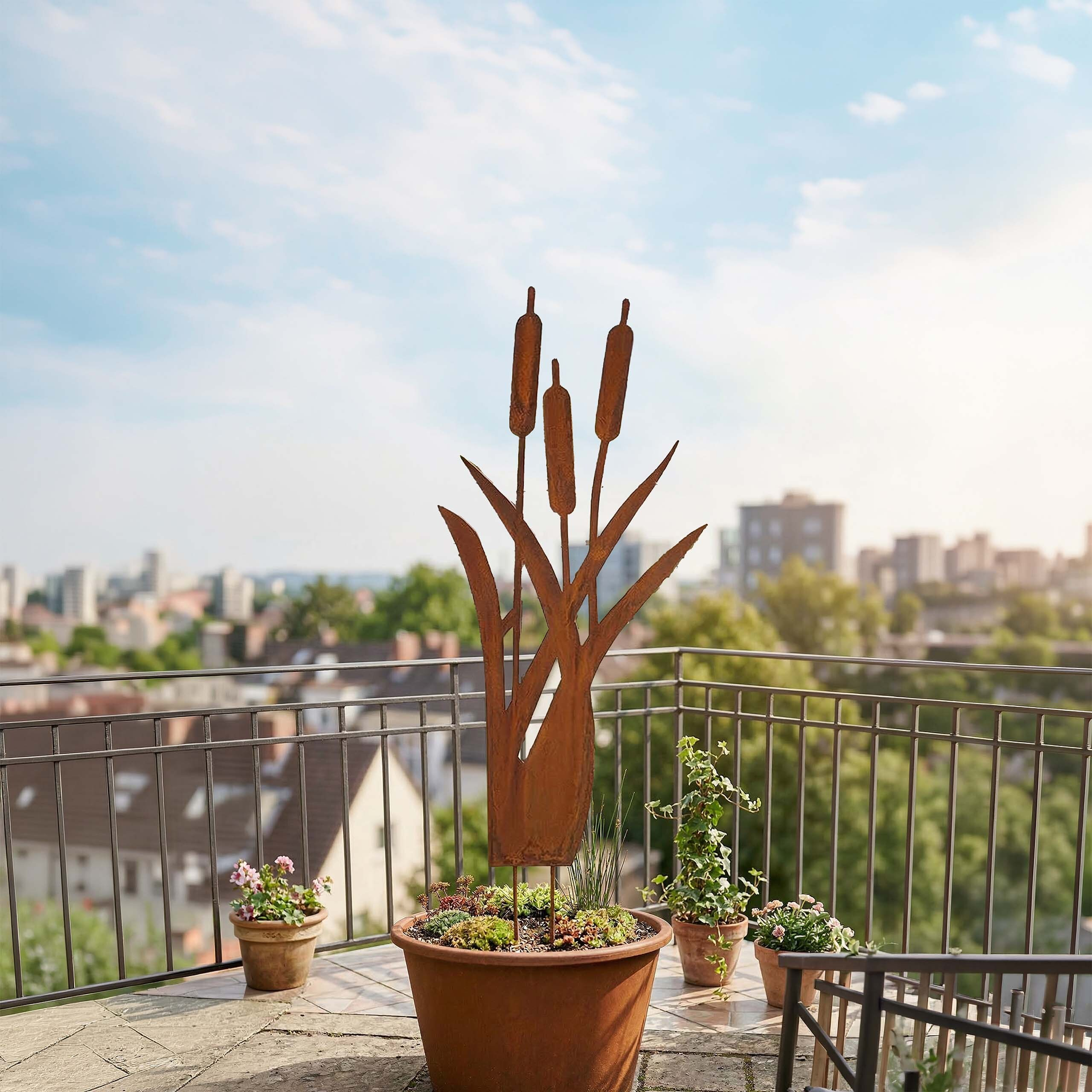 Eine rostige Metall-Rohrkolbenskulptur steht in einem großen Topf auf einem sonnigen Balkon mit Stadtblick.