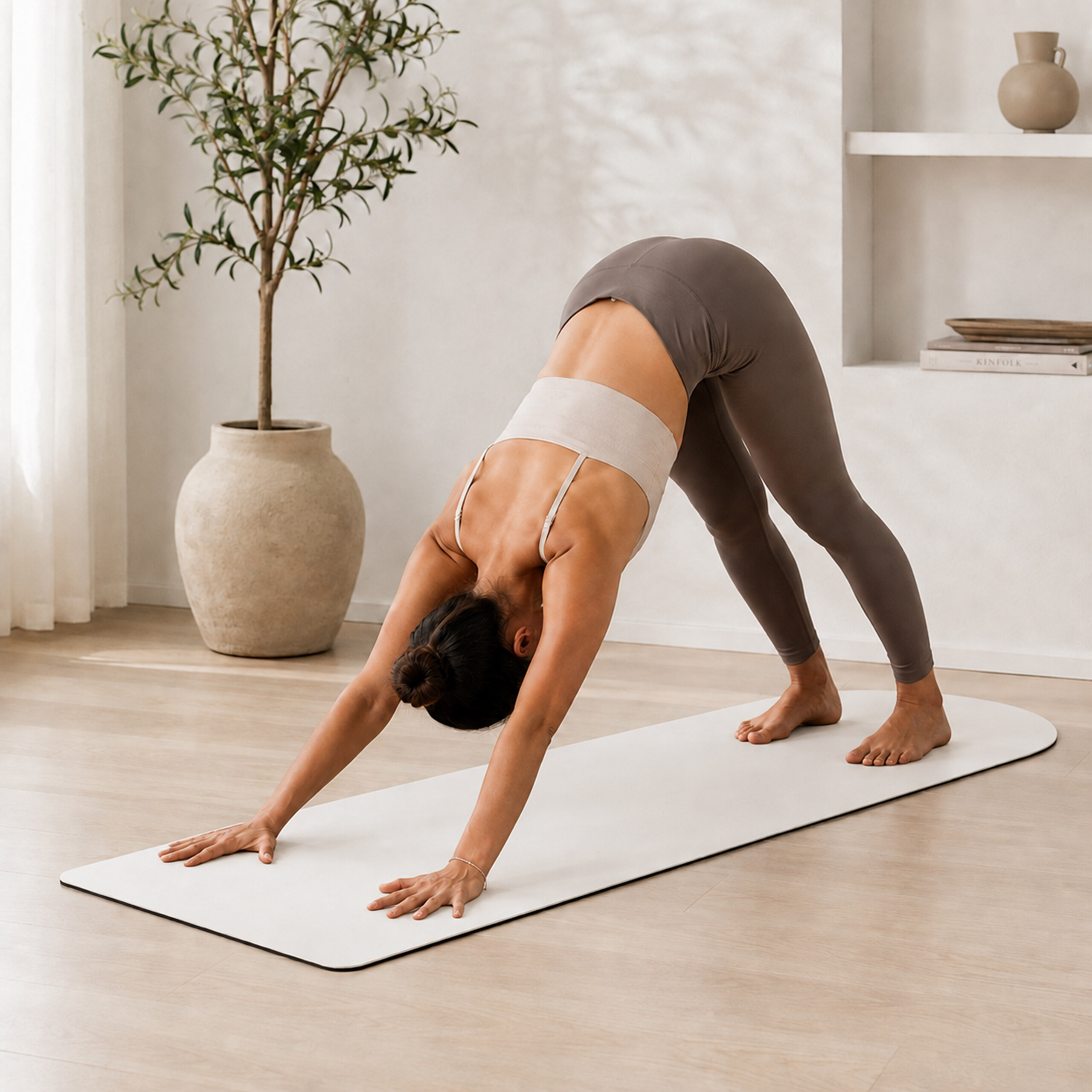 Eine Frau in grauen Leggings und einem beigen Oberteil praktiziert Yoga in der herabschauenden Hundeposition auf einer Matte.