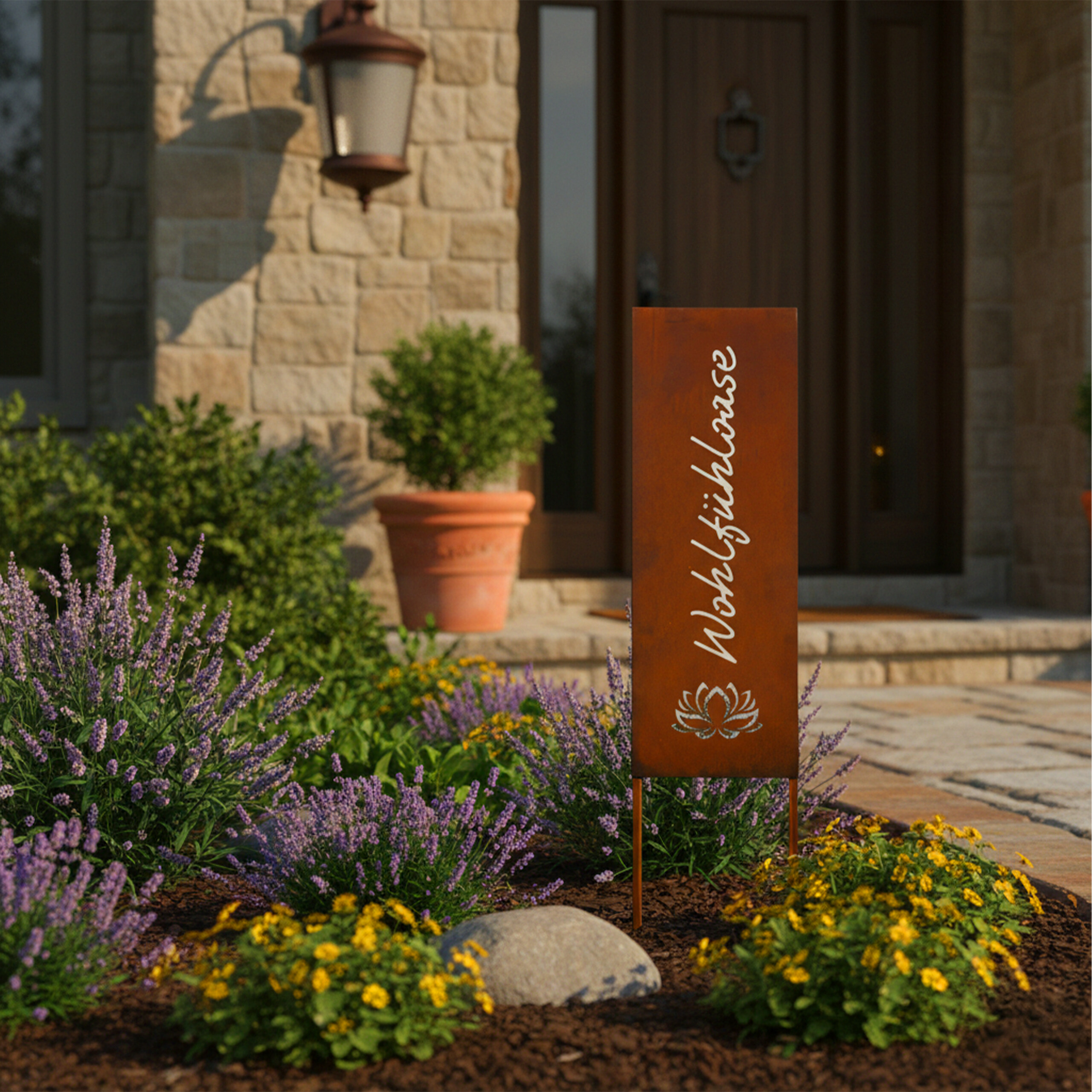 Rostiges Schild mit Aufschrift "Wohlfühloase" vor einem Haus mit Garten, Lavendel und gelben Blumen.