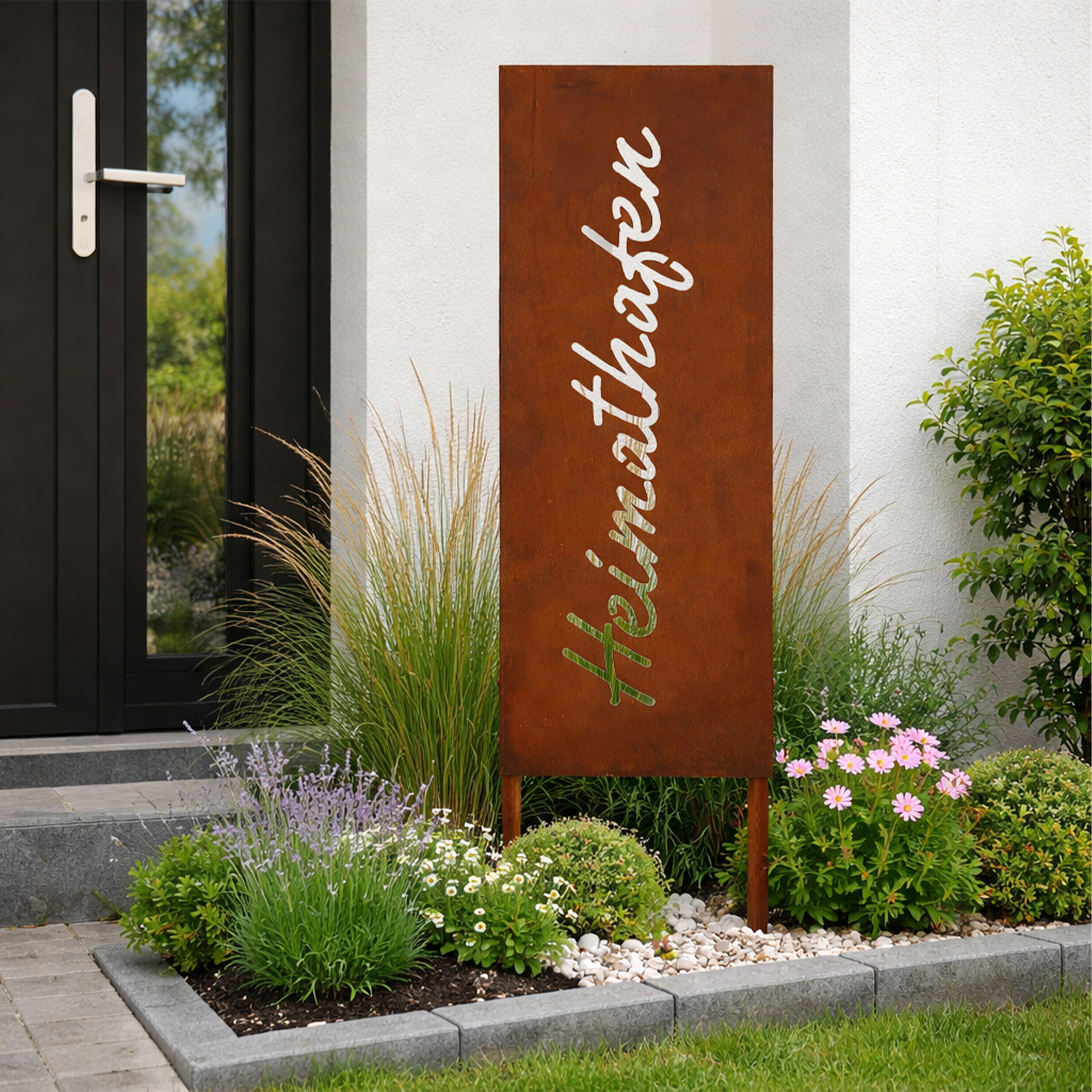 Ein rostiges Metallschild mit ausgeschnittenem Text steht in einem Gartenbeet mit Blumen und Gras neben einem weißen Haus.