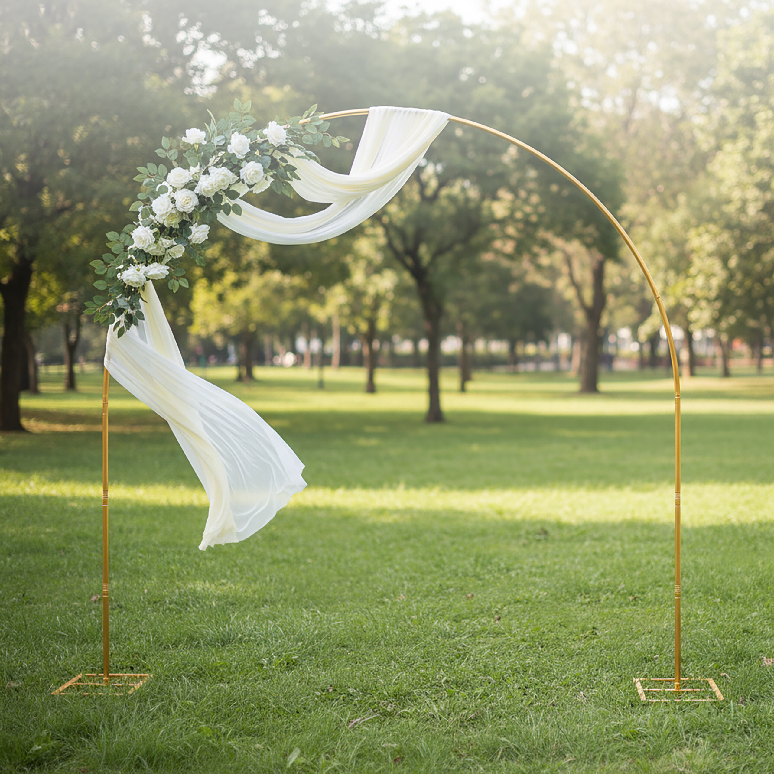 Ein goldener Hochzeitsbogen, verziert mit weißen Blumen und Stoff, steht in einem grünen Park.