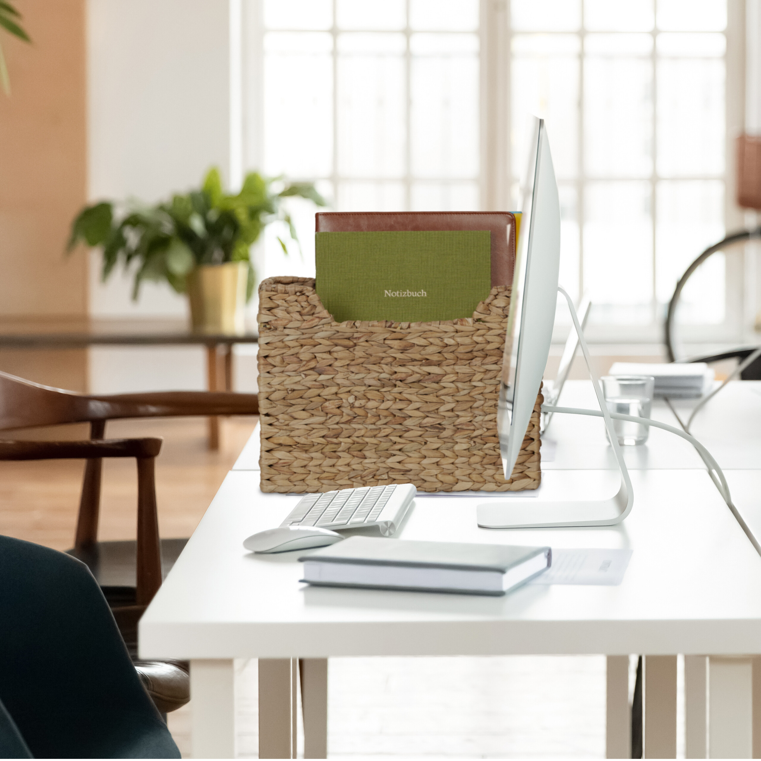 Einhelliger Schreibtisch mit Computer, Tastatur, Maus, Notizbüchern in einem Korb und einer Pflanze im Hintergrund.