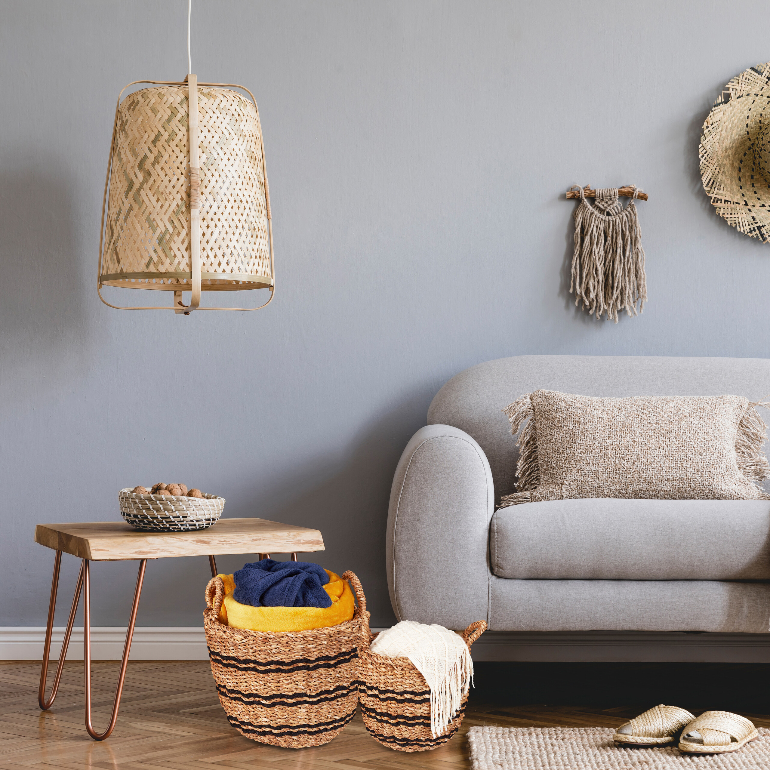 Ein gemütliches Wohnzimmer mit grauem Sofa, Holztisch, Weidenkörben, Lampe und Wanddeko an einer grauen Wand.