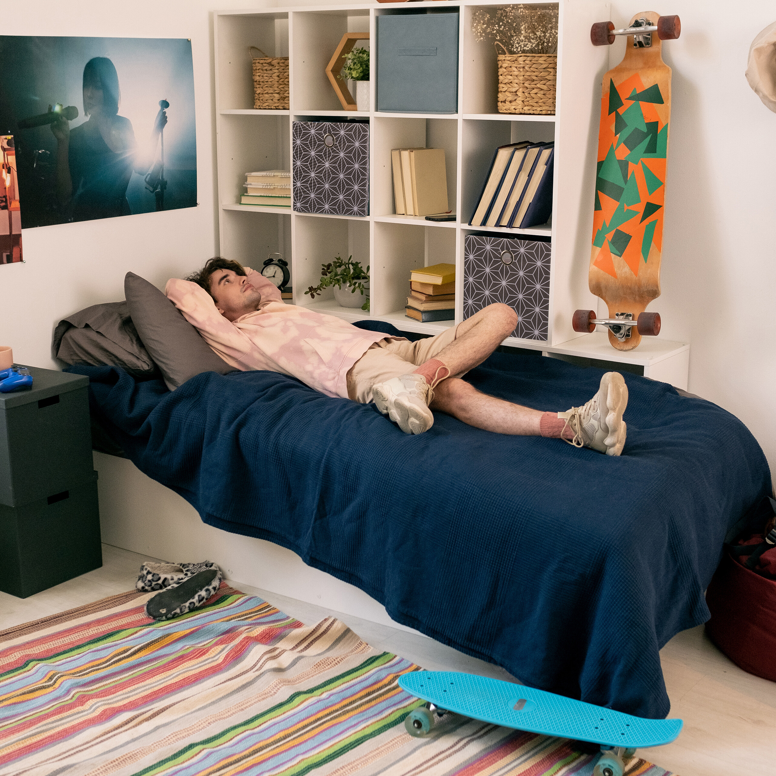 Ein junger Mann liegt auf einem blauen Bett in einem Zimmer mit Regalen, Skateboards und einem bunten Teppich.