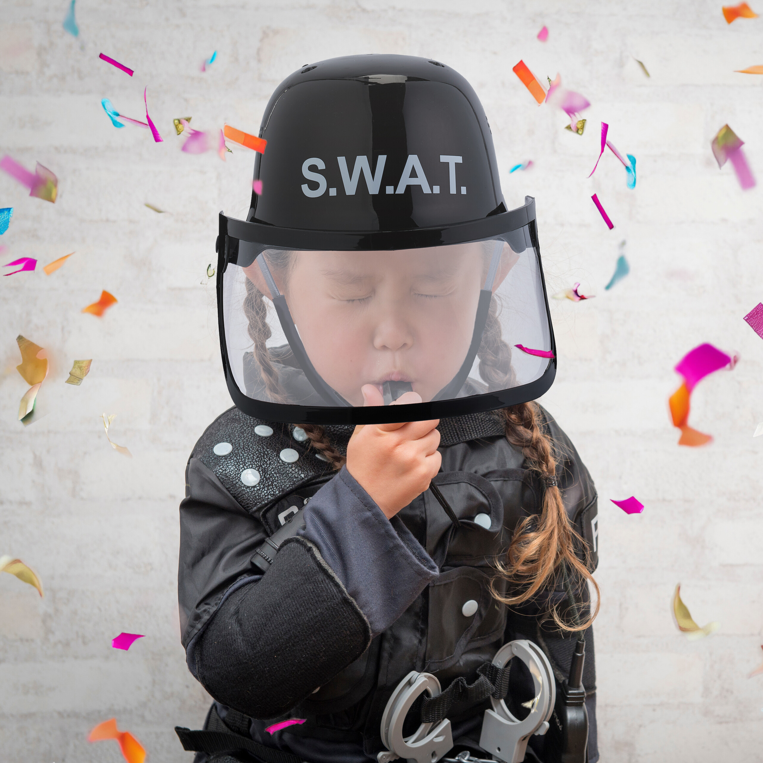 Ein Kind in SWAT-Ausrüstung und Helm bläst im Konfektionsregen in eine Pfeife vor einer weißen Wand.