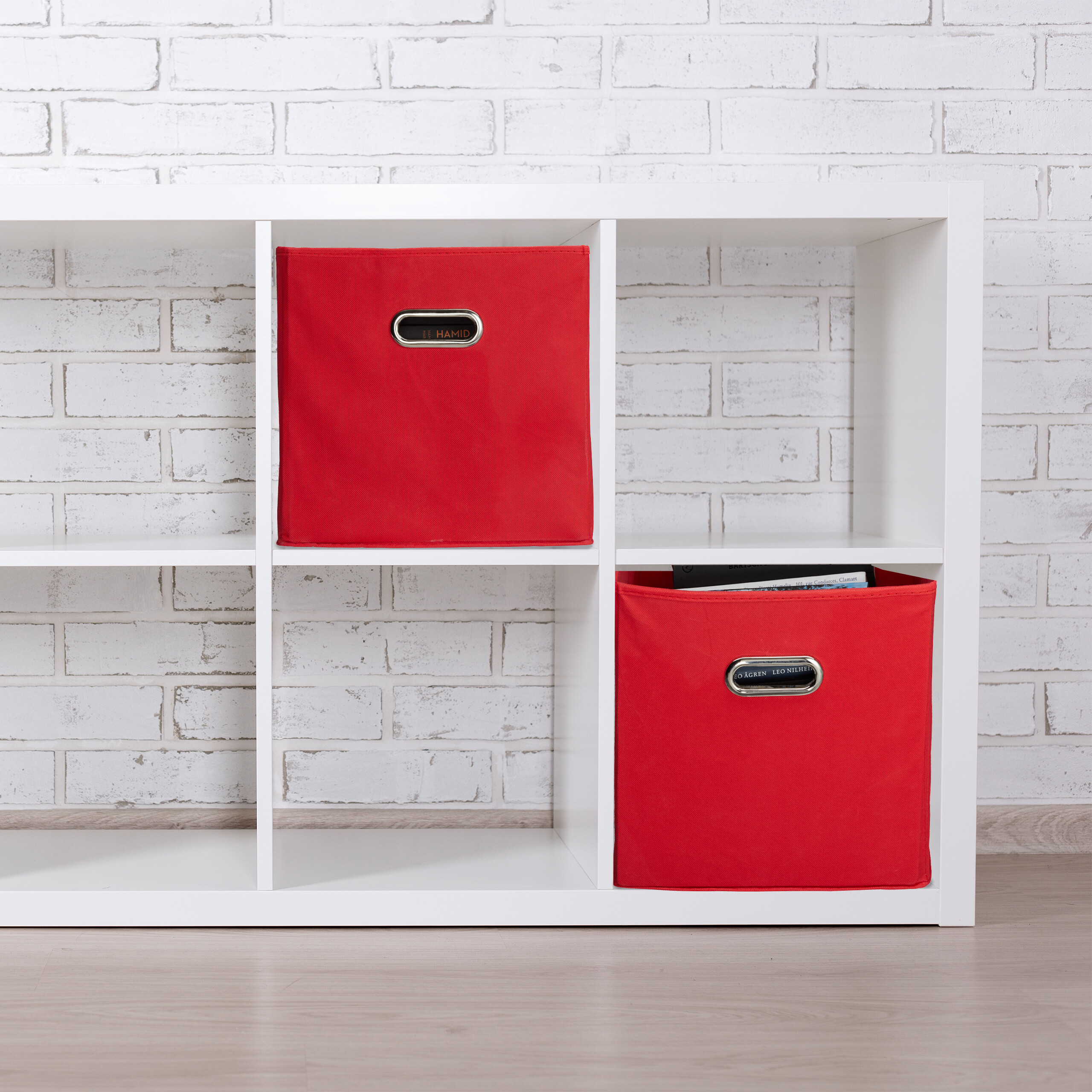Zwei rote Stoffboxen mit Metallgriffen stehen in einem weißen Regal vor einer hellen Backsteinwand.