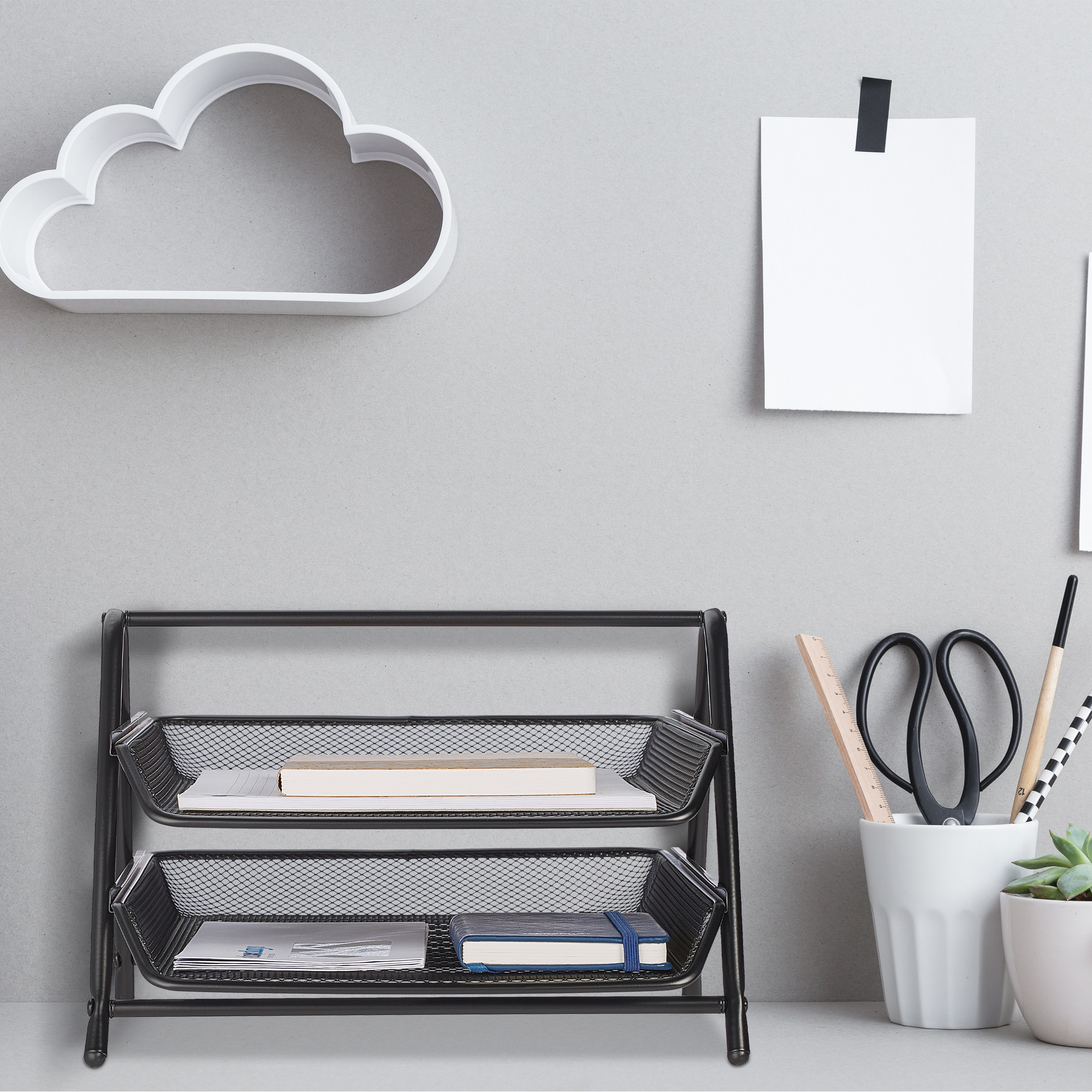 Ein Schreibtisch mit schwarzem Metallregal, Schreibwaren im Becher, Wolkenregal und Notizblatt an grauer Wand.