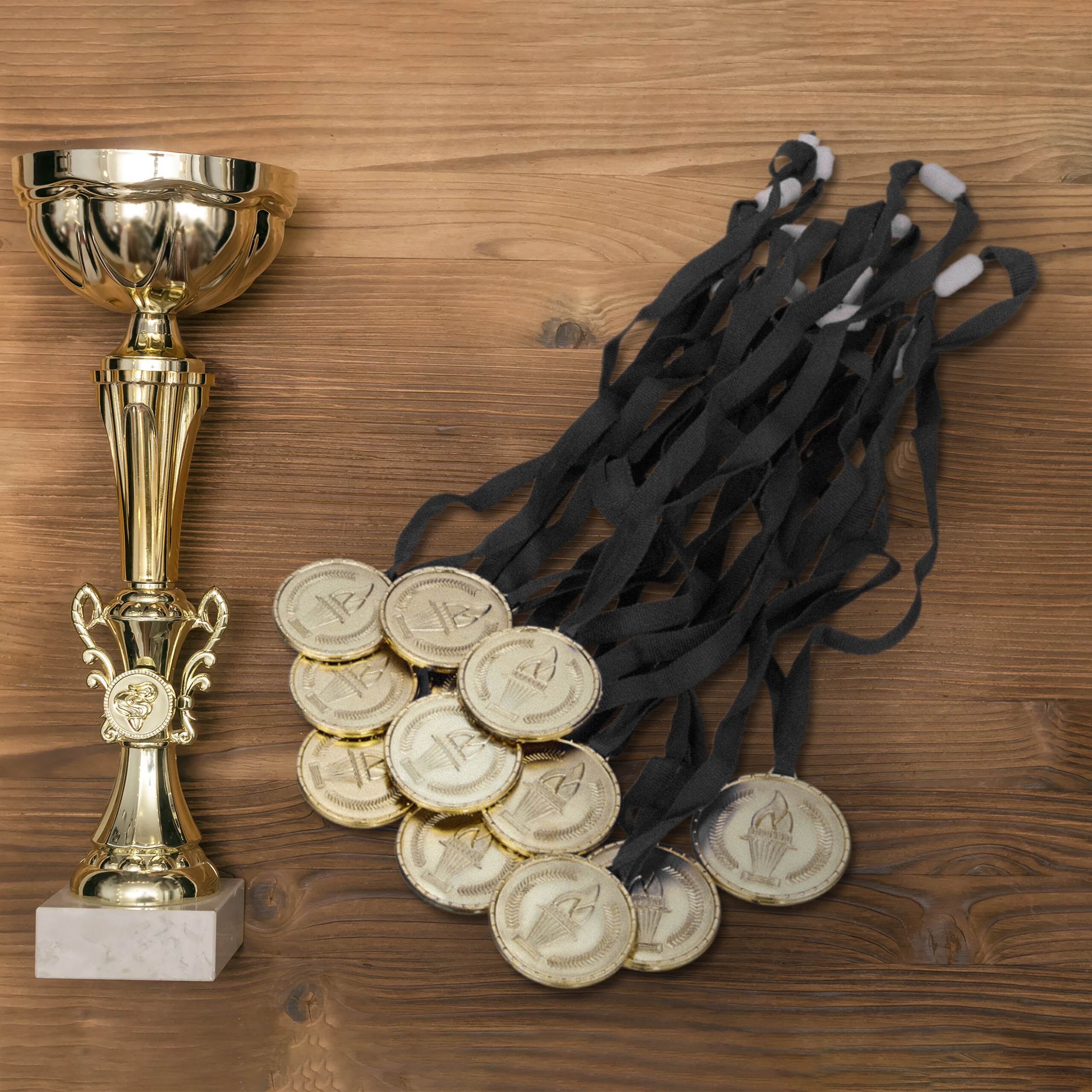 Ein goldener Pokal auf einem Sockel neben einem Haufen goldener Medaillen mit schwarzen Bändern auf Holz.