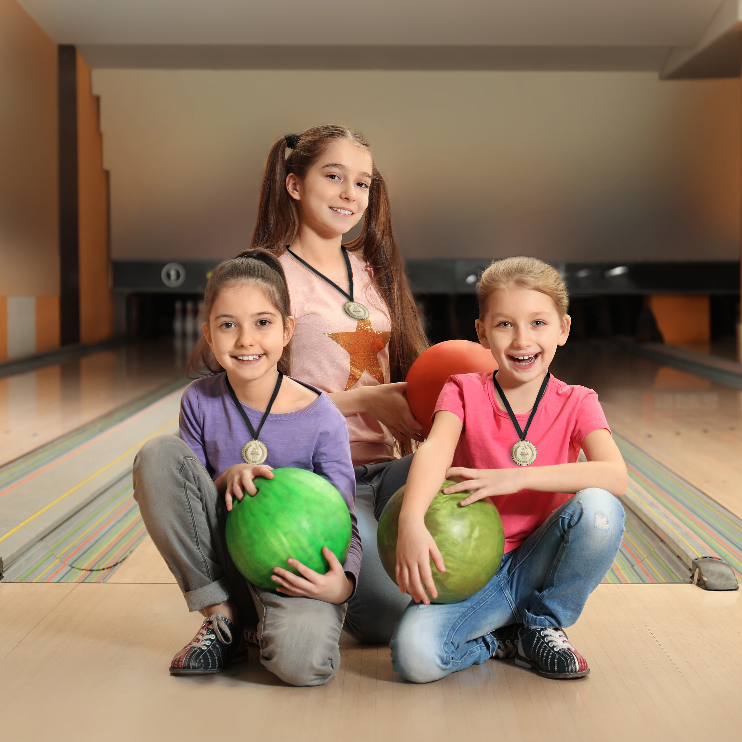 Drei glückliche Mädchen mit Medaillen posieren mit Bowlingkugeln auf einer Bowlingbahn.