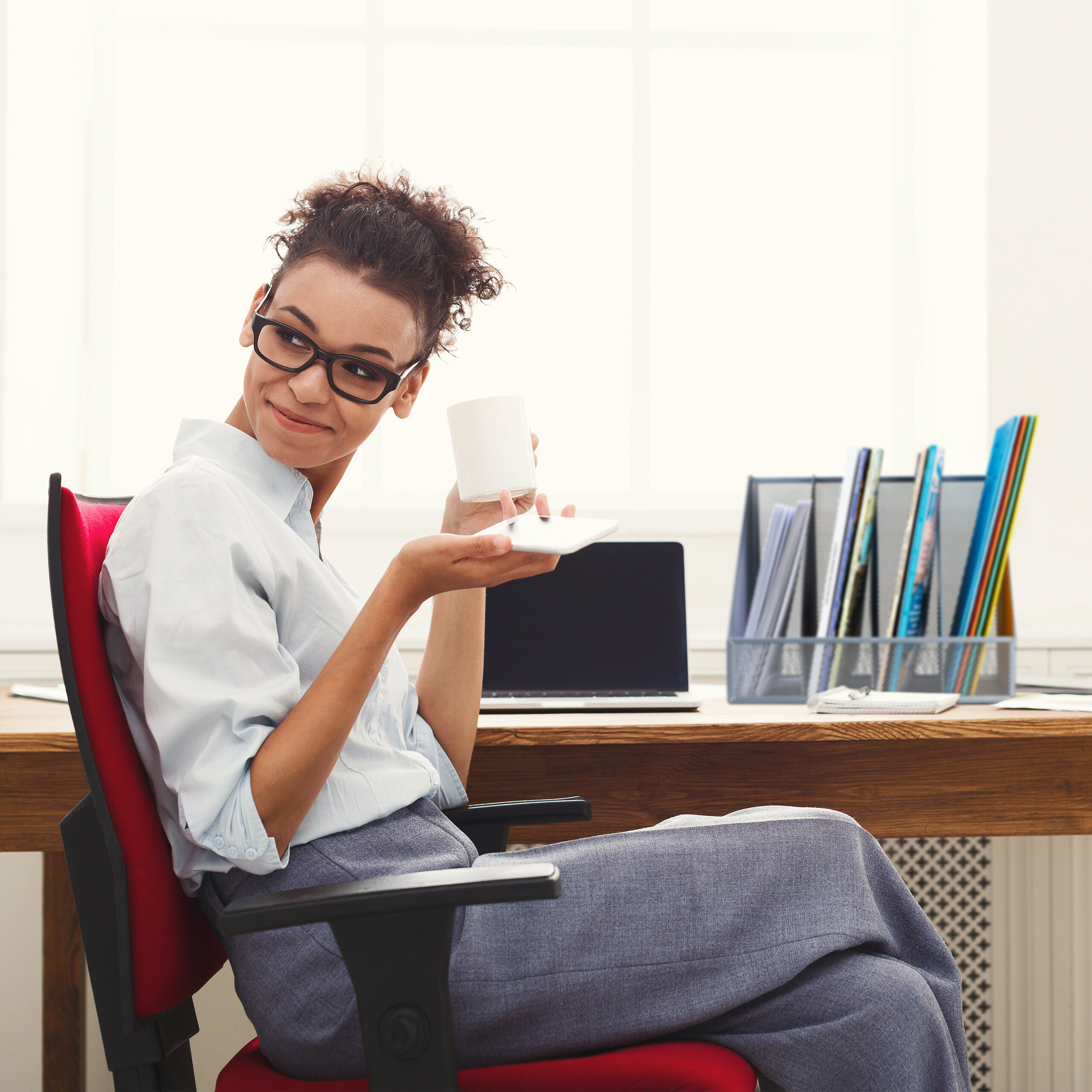 Eine smiling Frau mit Brille sitzt in einem roten Bürostuhl, hält eine Tasse und schaut zur Seite.