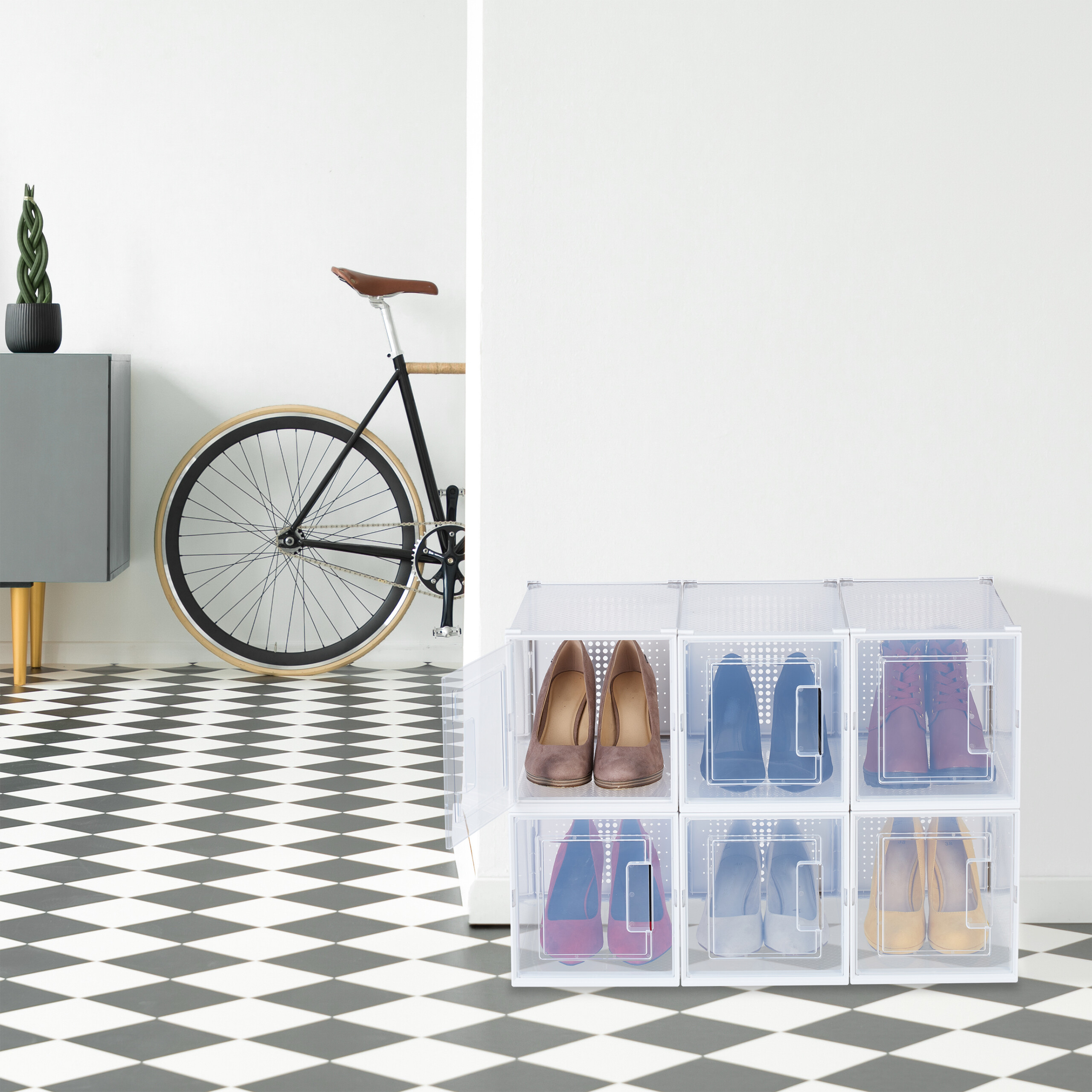 Transparente Schuhkartons stehen auf einem schwarz-weiß gefliesten Boden neben einem Fahrrad und einer grauen Kommode.