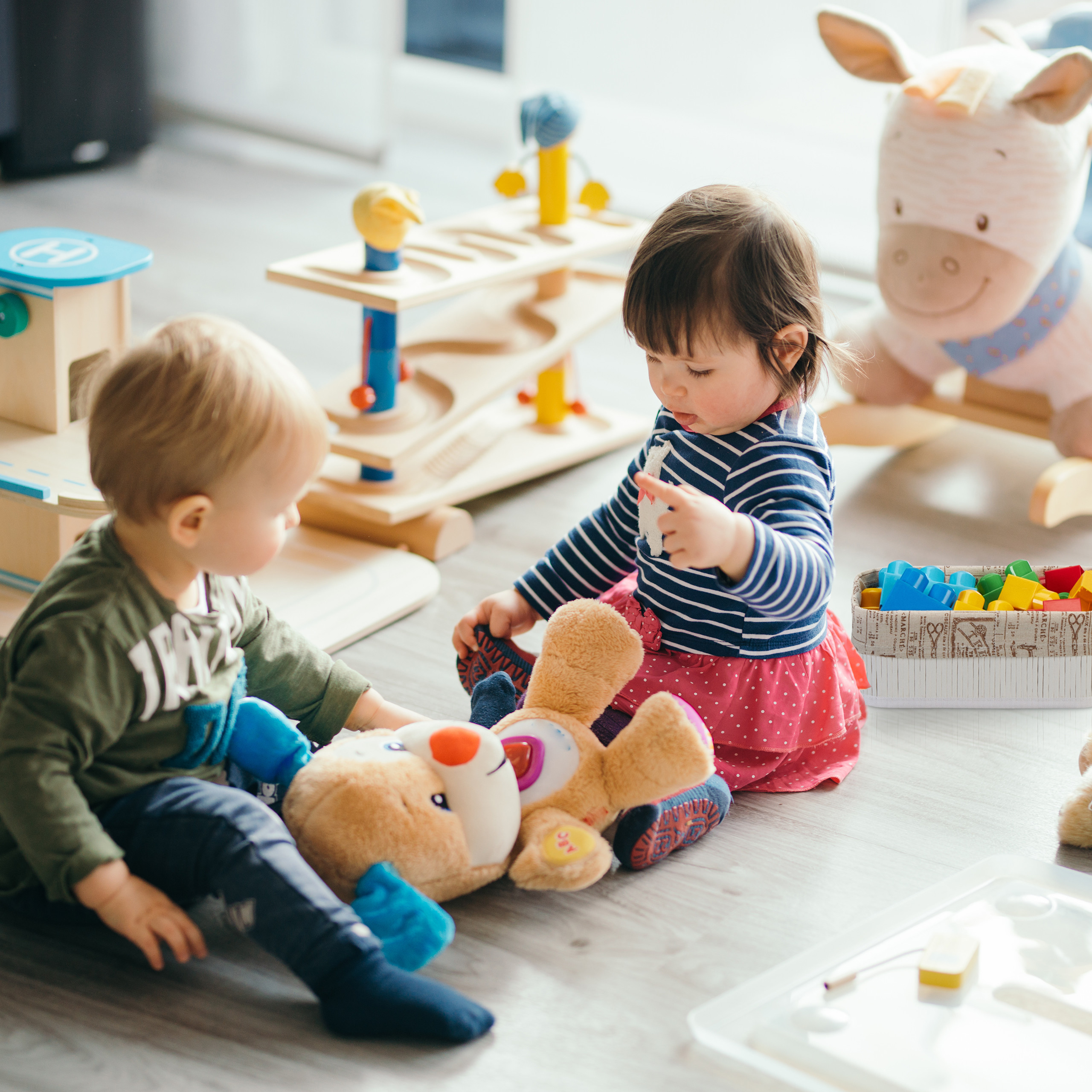Zwei Kleinkinder sitzen auf einem Holzboden und spielen mit Plüschtieren und Holzspielzeug.