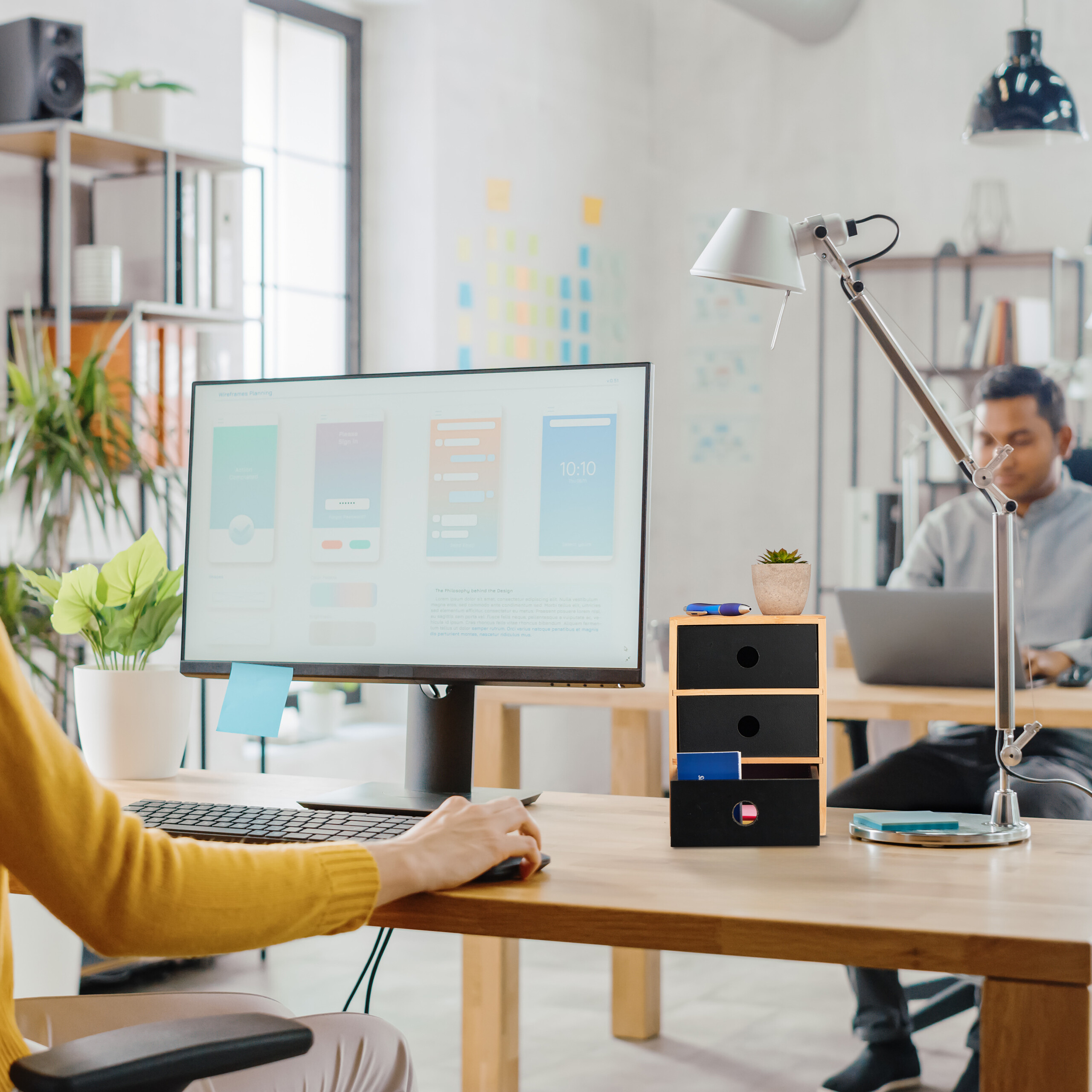 Designer arbeitet im Büro am Computer mit App-Design, ein Kollege sitzt im Hintergrund.