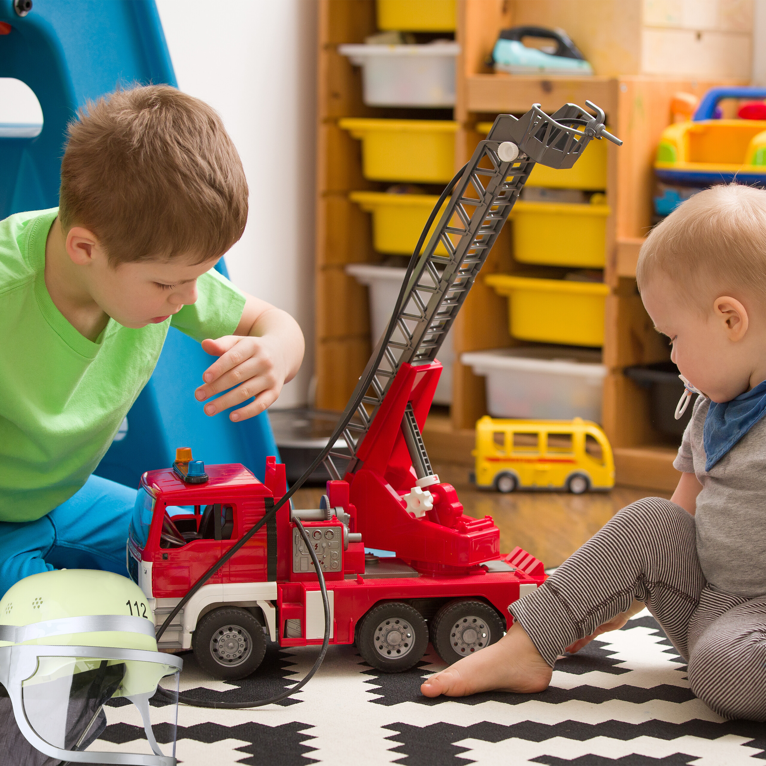 Zwei Jungen spielen in einem Spielzimmer auf einem gemusterten Teppich mit einem großen roten Spielzeug-Feuerwehrauto.