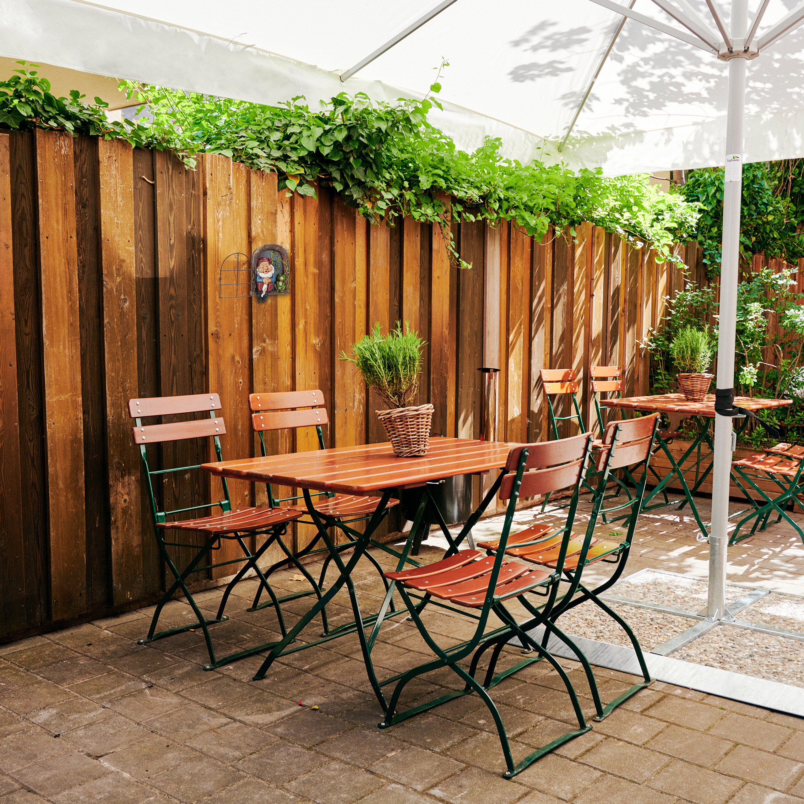 Holztische und Stühle mit grünem Metallgestell im Biergarten, im Hintergrund ein Holzzaun mit Pflanzen.