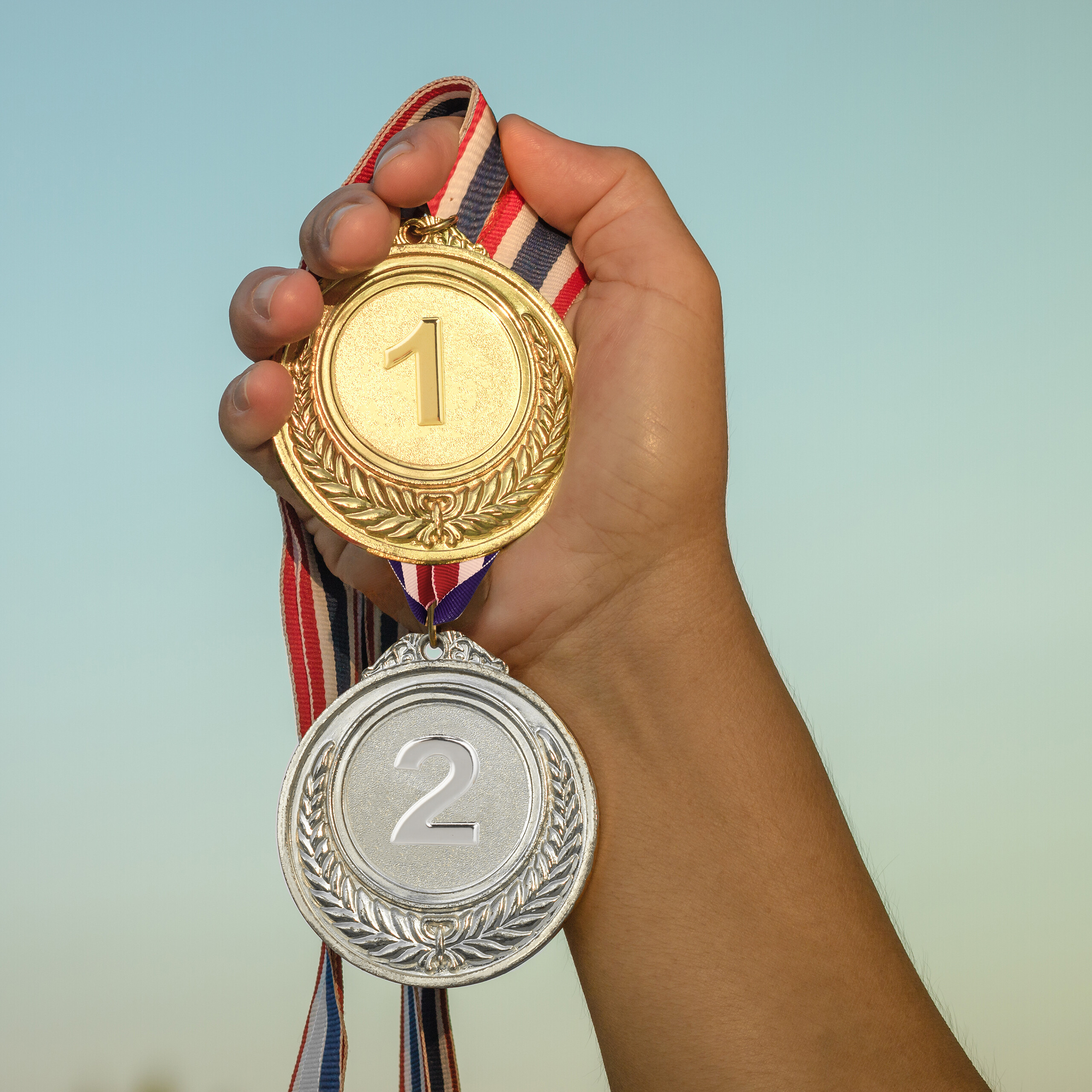 Eine Hand hält eine goldene Medaille mit einer Eins und eine silberne Medaille mit einer Zwei an bunten Bändern.
