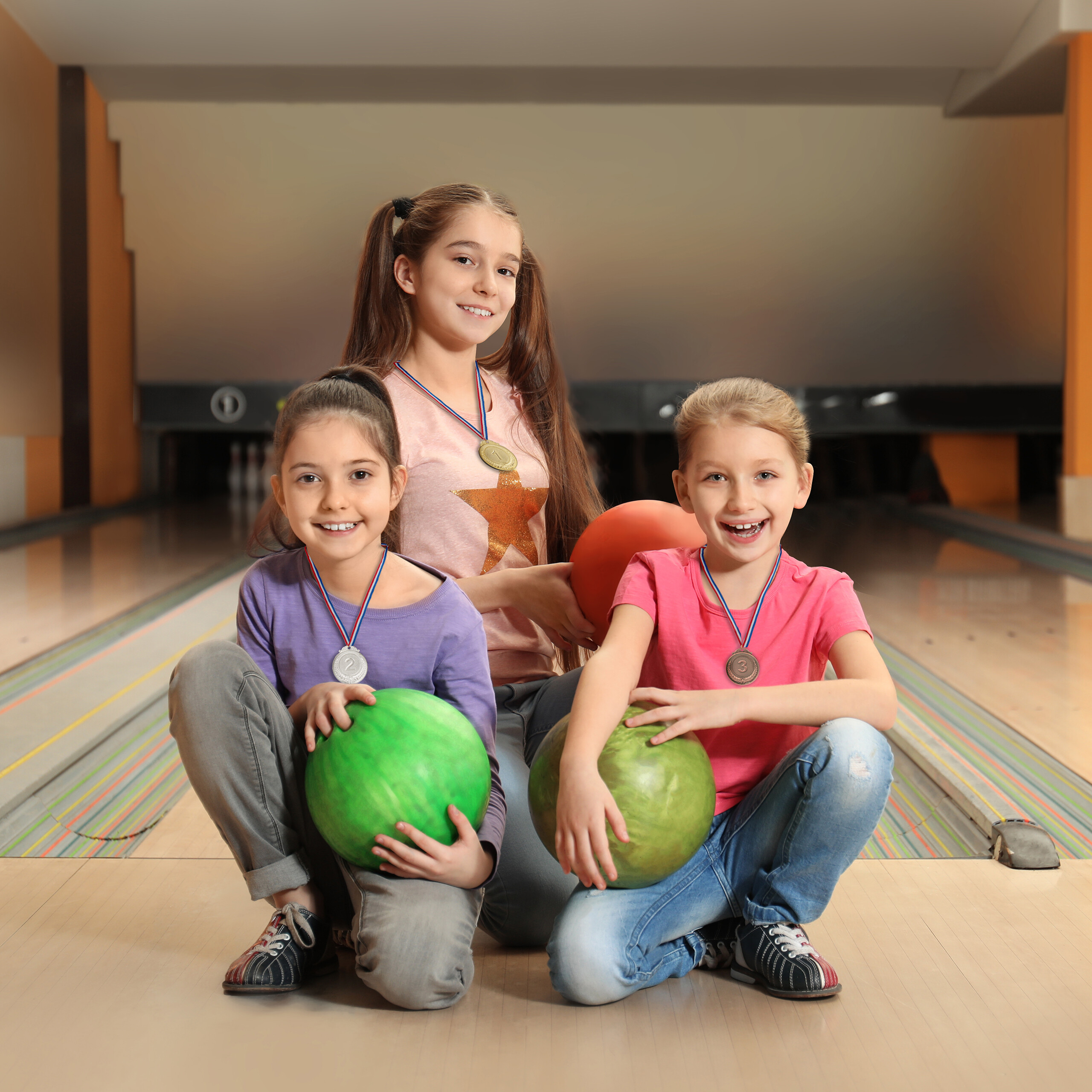 Drei glückliche Mädchen sitzen mit Medaillen und Bowlingkugeln auf einer Bowlingbahn.
