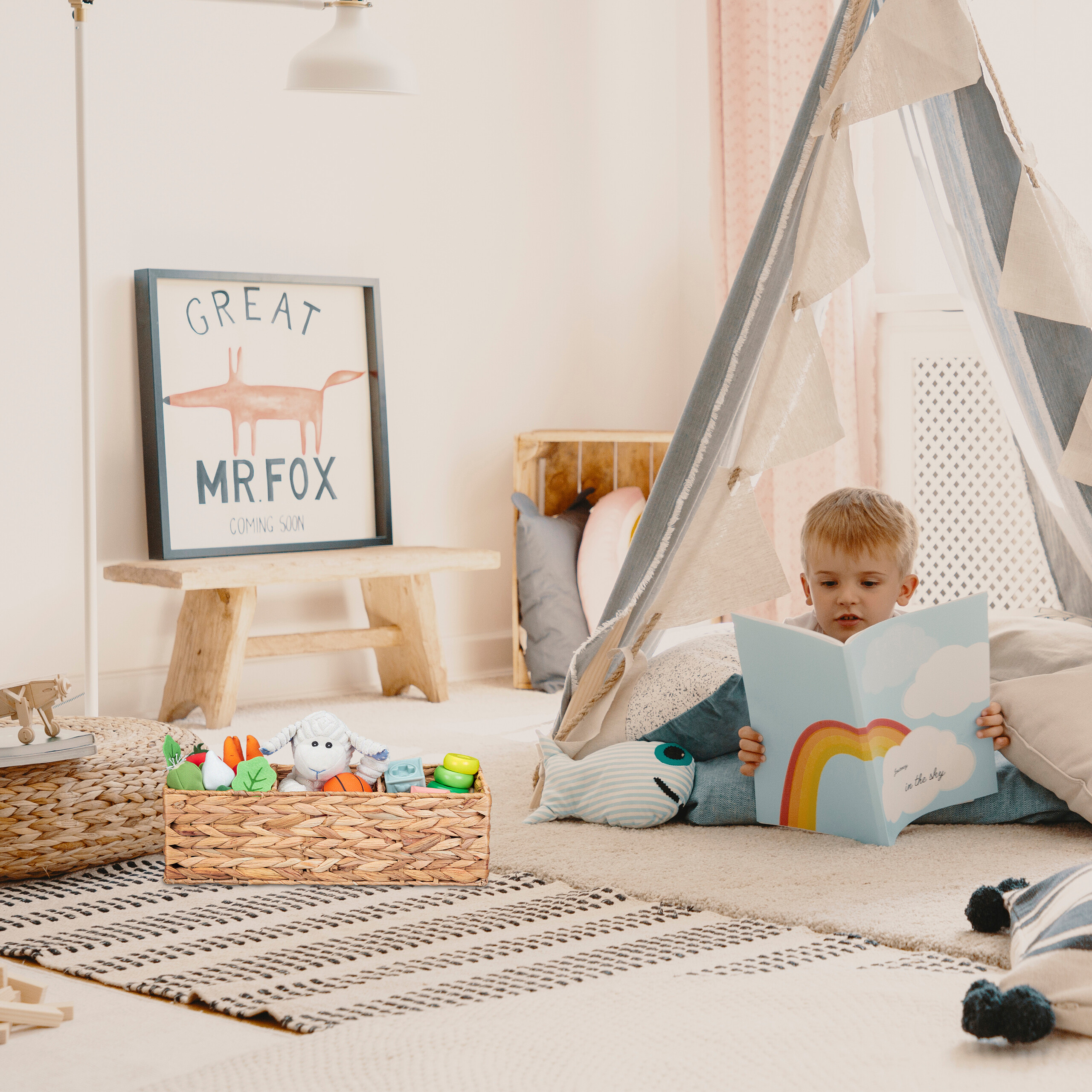 Ein kleiner Junge liest in einem Tipi-Zelt ein blaues Buch mit Regenbogen, umgeben von Spielzeug.