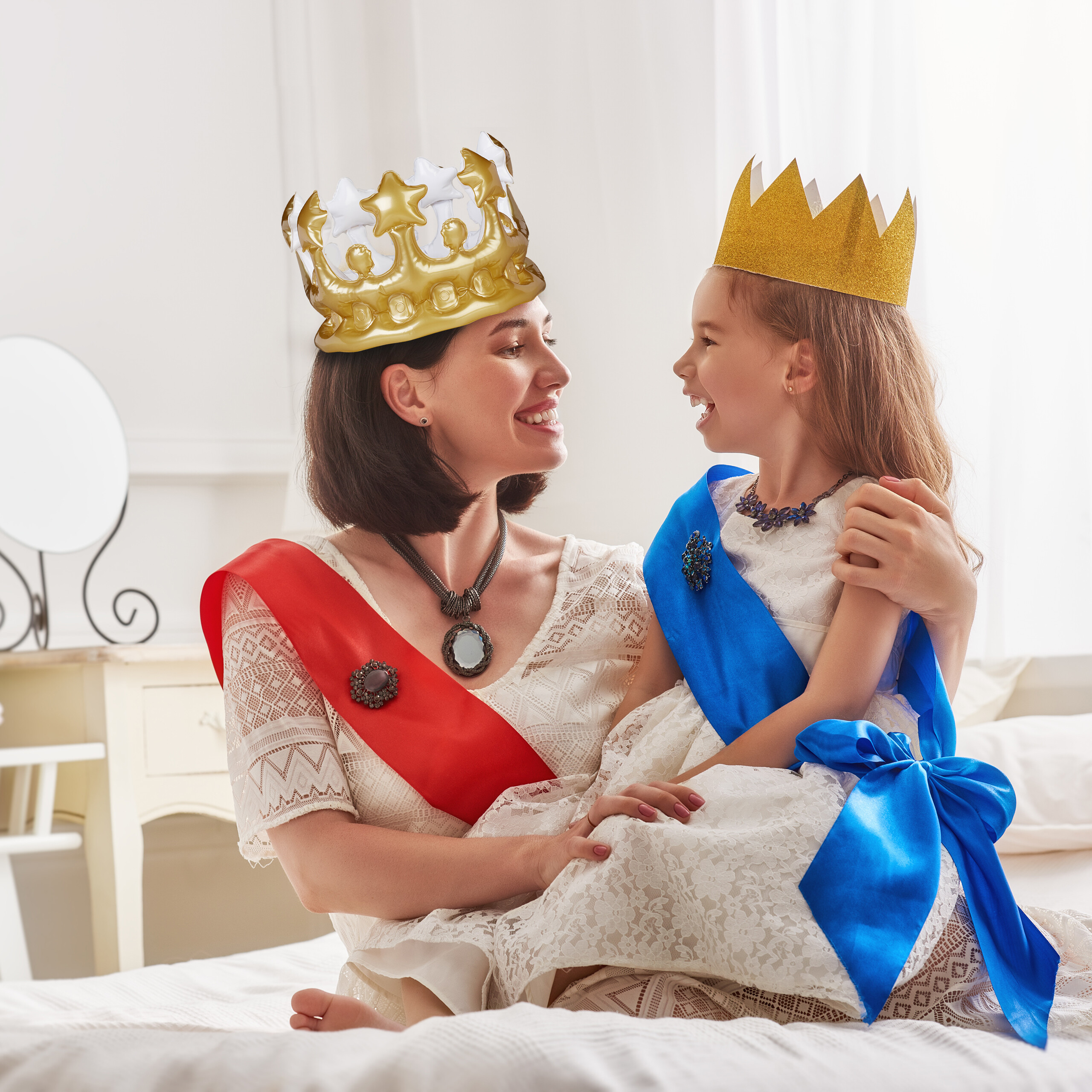 Eine Frau in weißem Kleid und großer Goldkrone sitzt mit ihrer kleinen Tochter, die eine Goldkrone trägt, auf dem Bett.