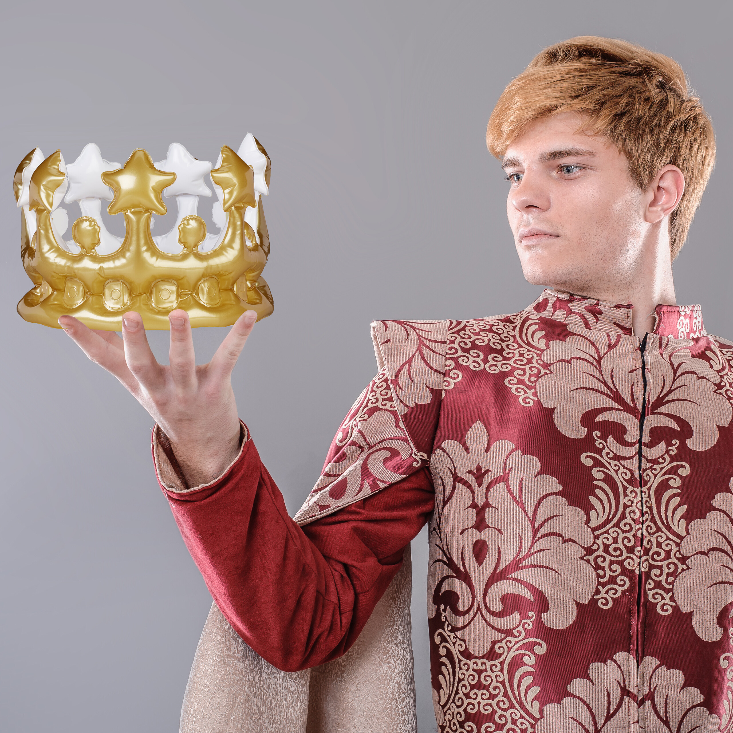Ein blondhaariger Mann in rot-goldener Renaissance-Kleidung hält eine goldene Krone in seiner offenen Hand.