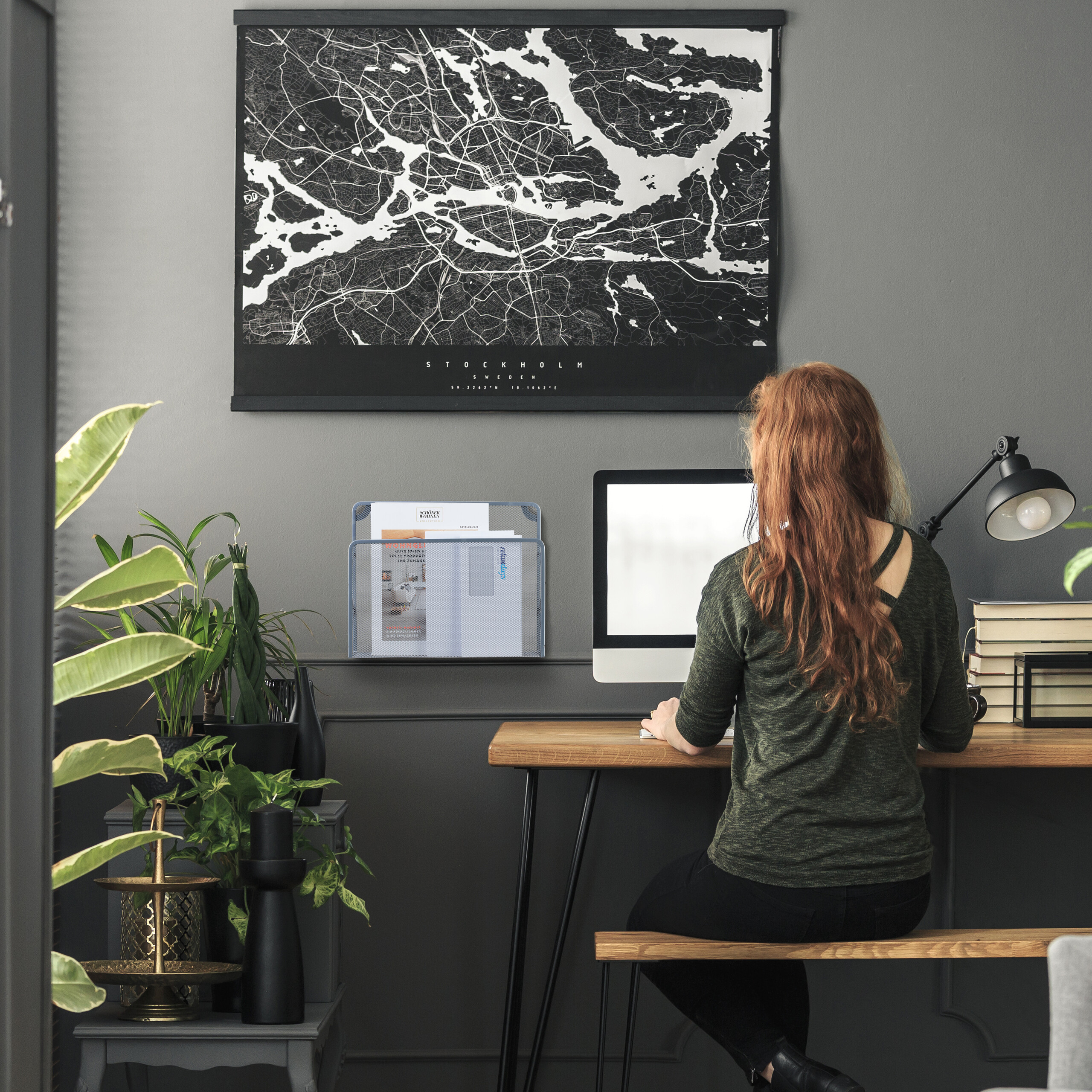 Eine rothaarige Frau sitzt an einem Holztisch und arbeitet am Computer, unter einer großen schwarz-weißen Landkarte.