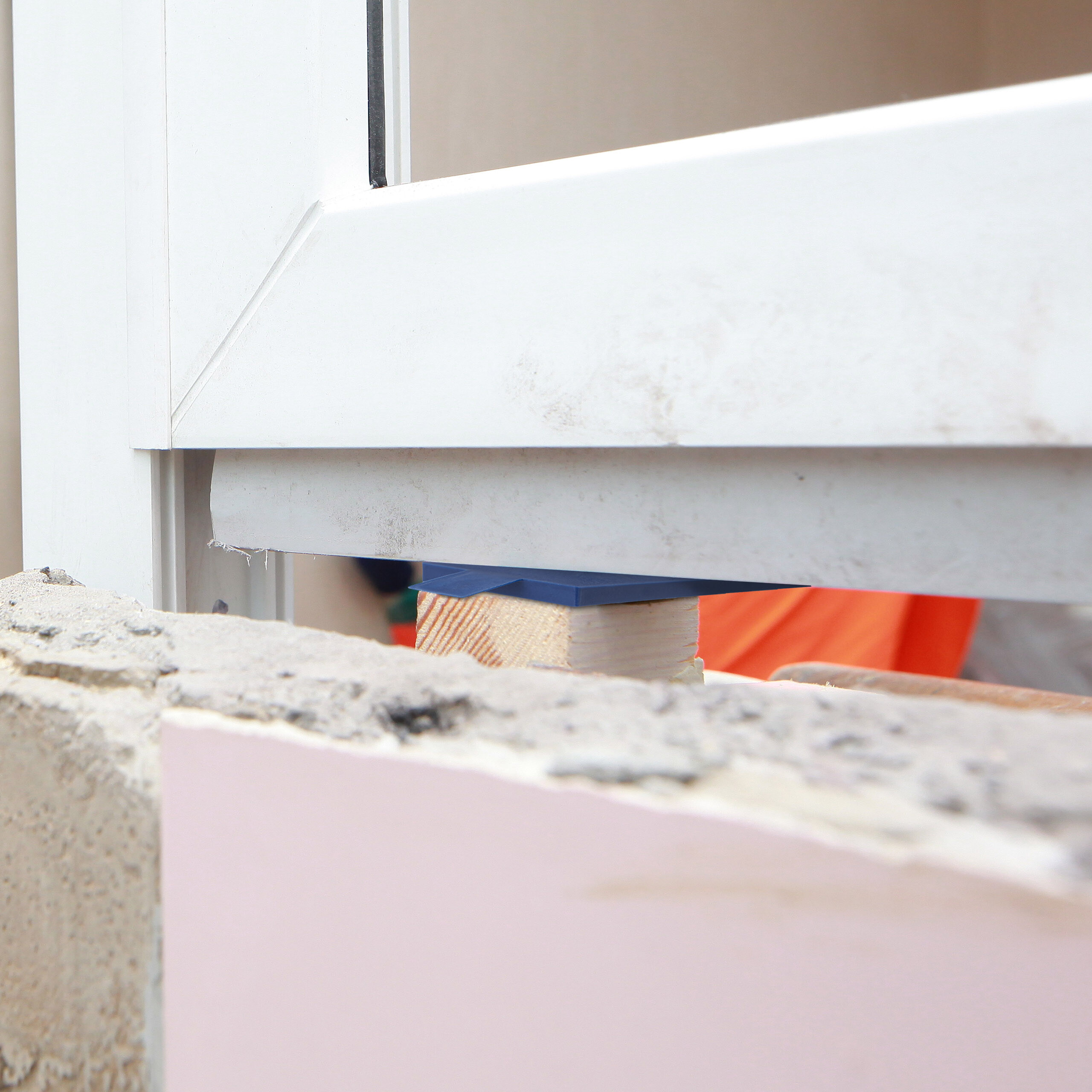 Ein weißer Fensterrahmen wird mit einem Holzklotz und einem blauen Keil auf einer Betonwand nivelliert.