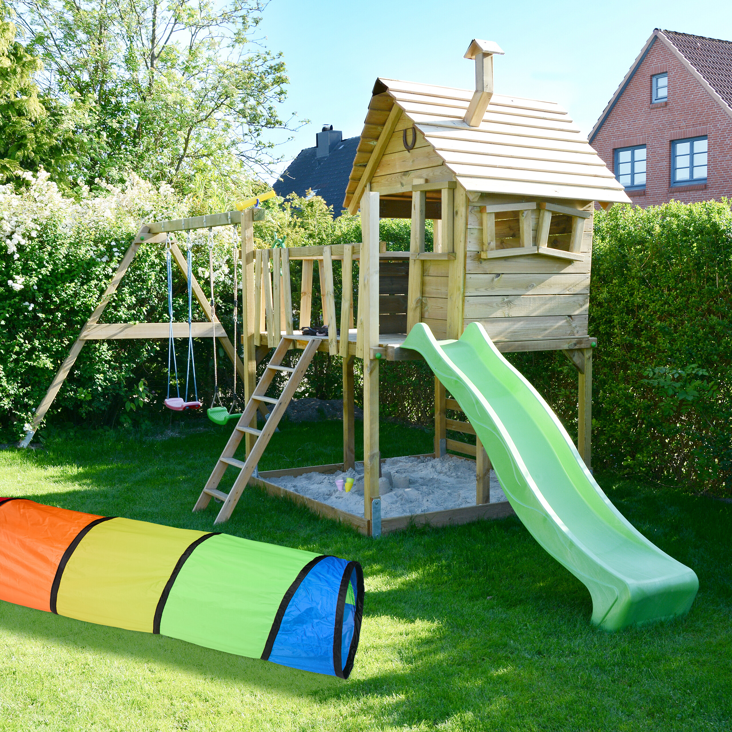 Holzspielplatz mit Rutsche, Schaukel und Sandkasten im Garten, davor ein bunter Stofftunnel.
