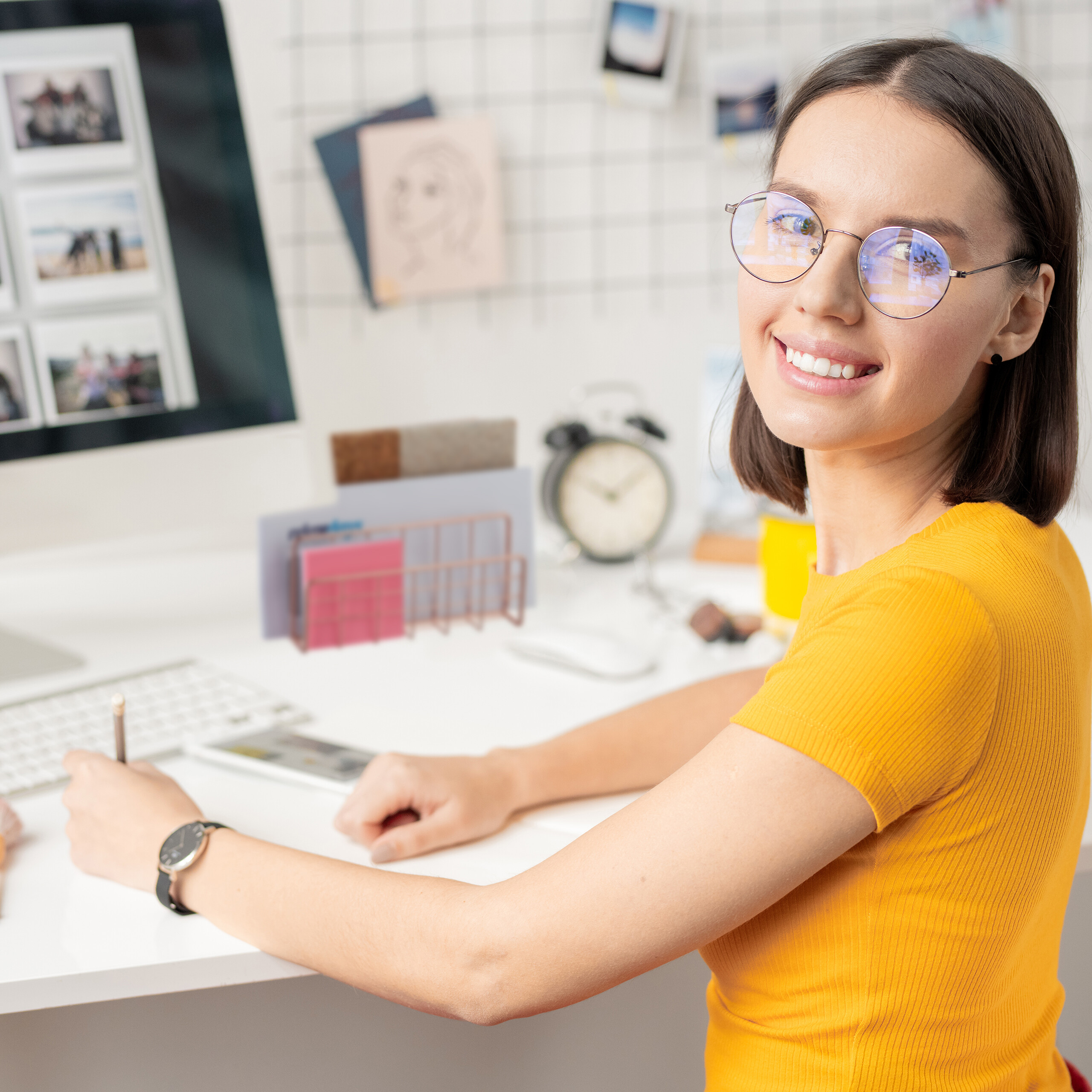 Eine lächelnde Frau in gelbem Shirt und Brille sitzt an ihrem weißen Schreibtisch und arbeitet am Computer.