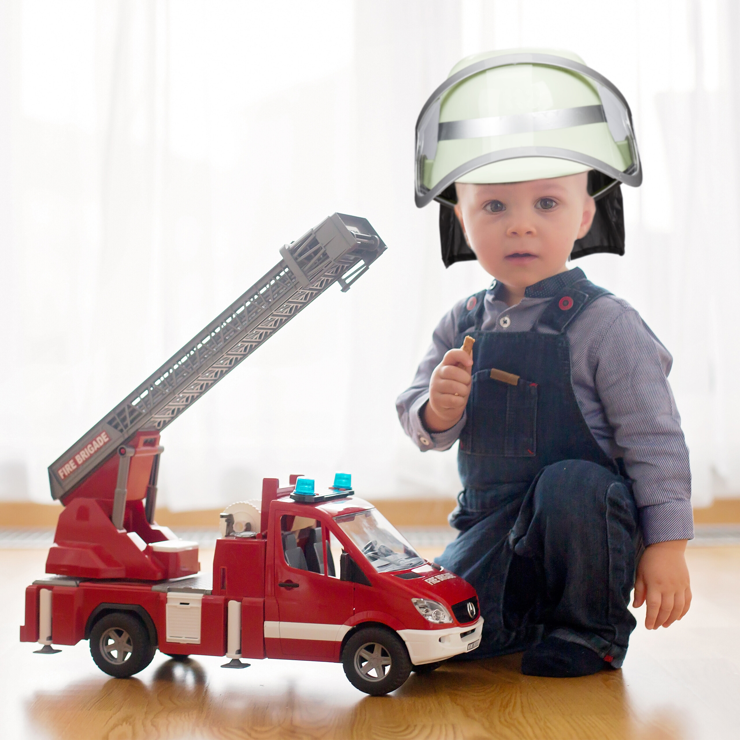 Ein Kleinkind in Latzhose und Feuerwehrhelm spielt auf einem Holzboden mit einem großen roten Spielzeug-Feuerwehrauto.