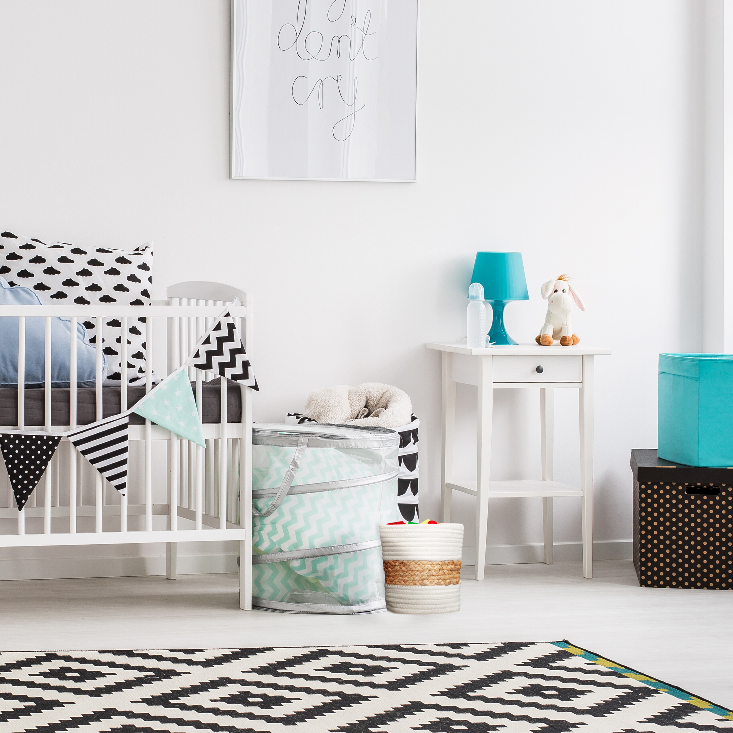 Ein weißes Babybett mit Kissen, eine Banderole, ein Nachttisch mit Lampe und Spielzeug, Teppich und Kisten in einem Zimmer.
