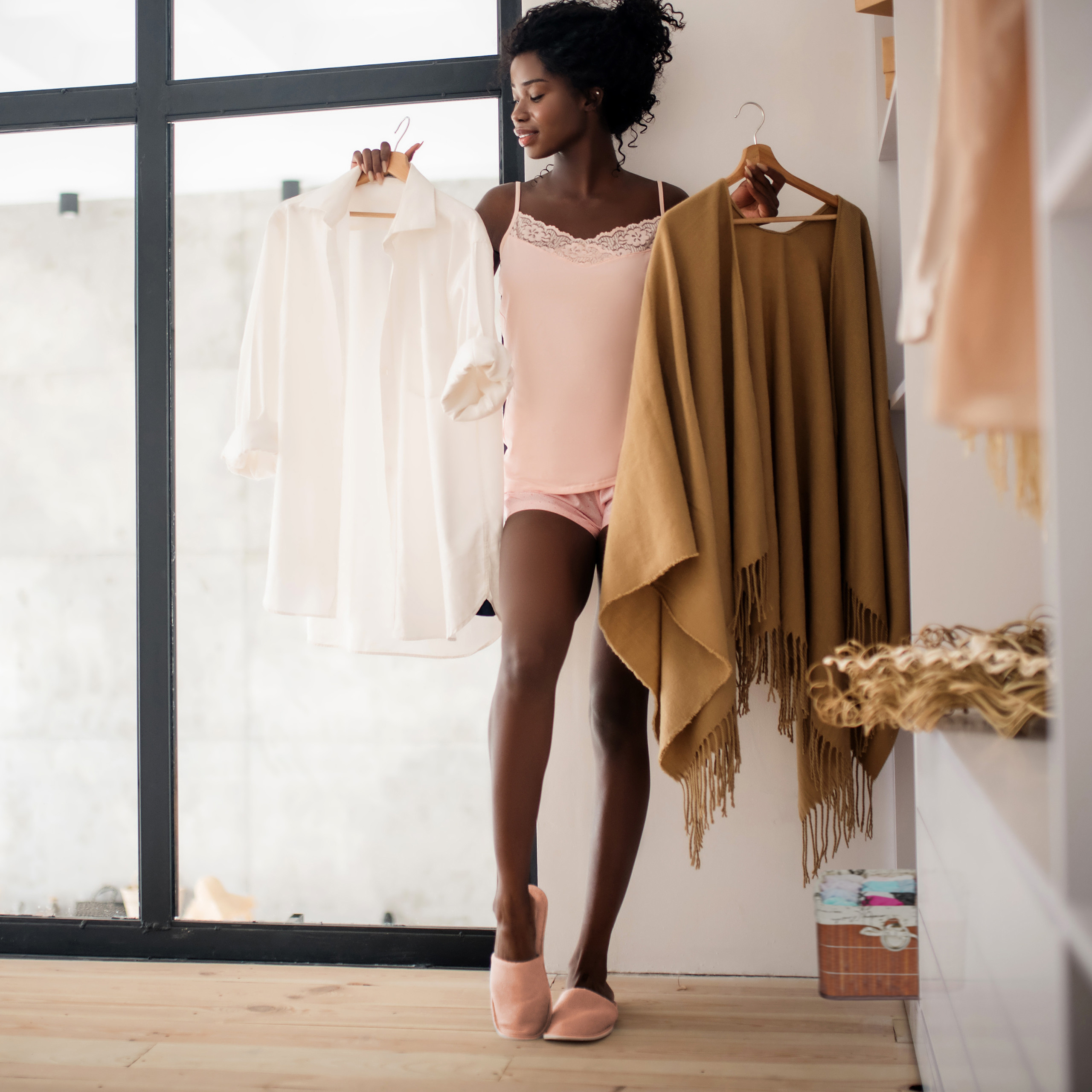 Eine Frau in rosa Pyjama hält in ihrem Kleiderschrank eine weiße Bluse und einen braunen Schal in den Händen.