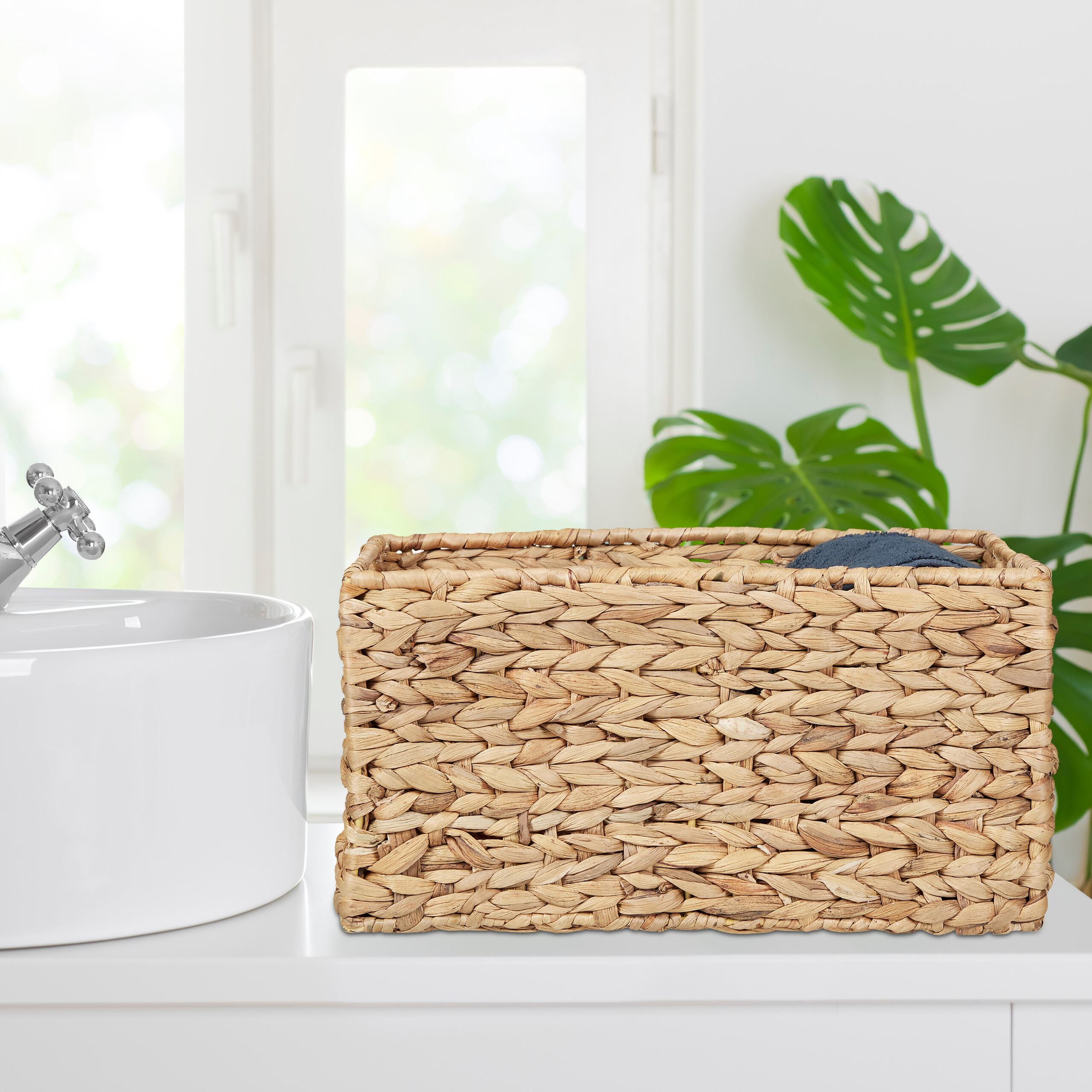 Ein geflochtener Weidenkorb steht neben einem Waschbecken auf einer weißen Theke vor einem hellen Fenster mit grüner Pflanze.
