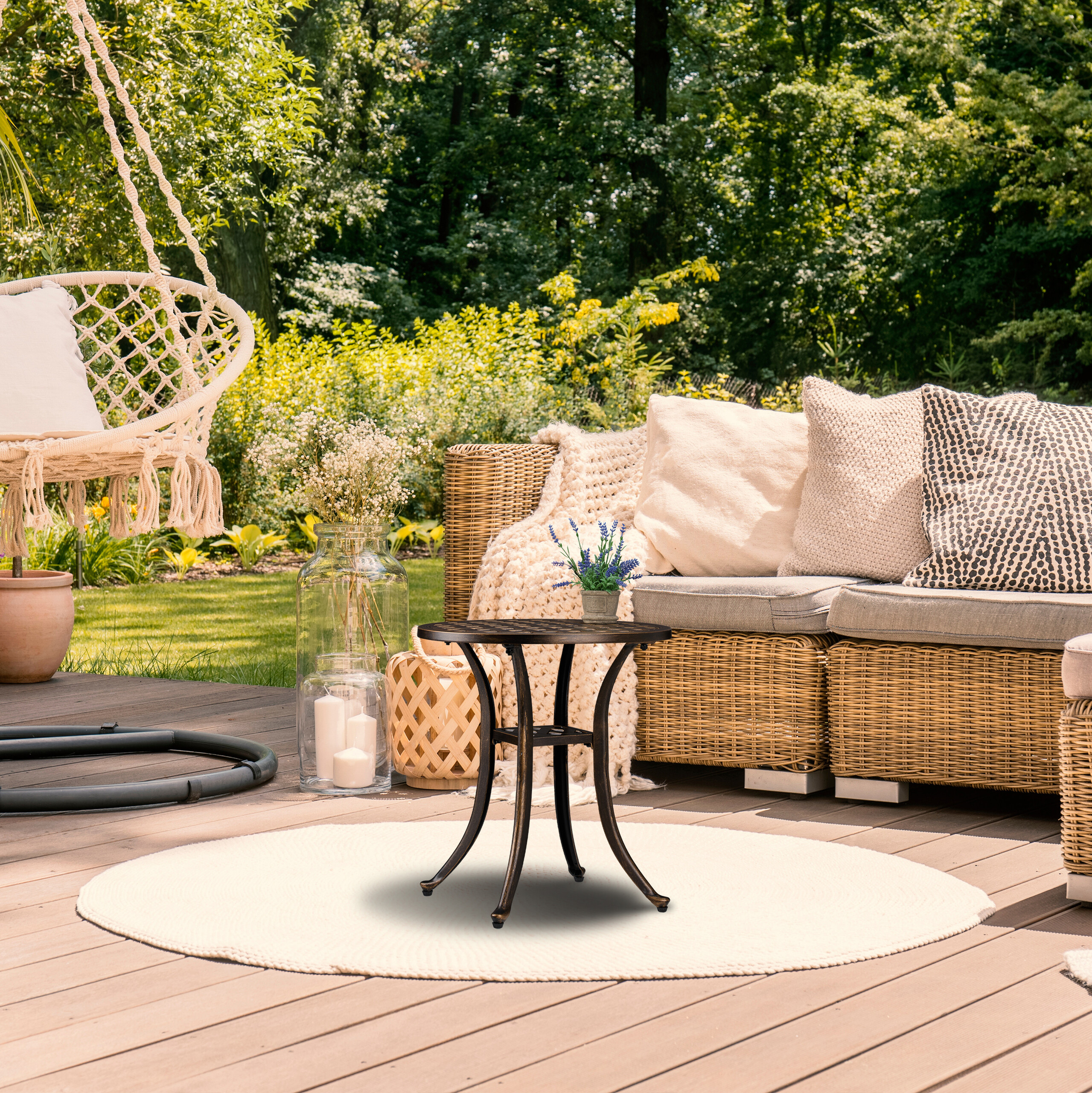 Ein runder Metalltisch auf einer Holzterrasse im Garten, mit einem Korbsofa, Blumen und einem Hängesessel im Hintergrund.