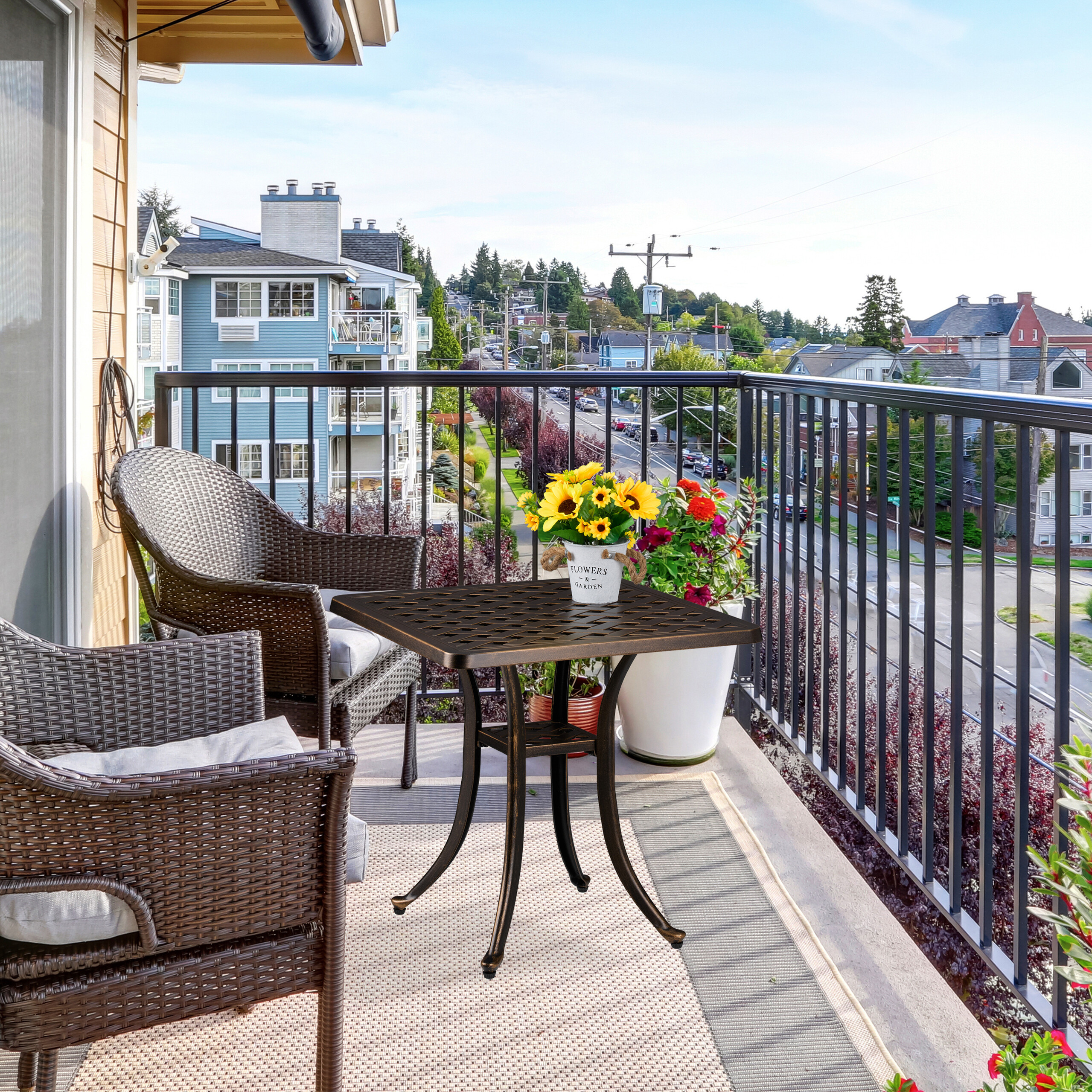 Ein sonniger Balkon hat einen kleinen dunklen Tisch, zwei Korbsessel und Blumen mit Blick auf eine Straße.