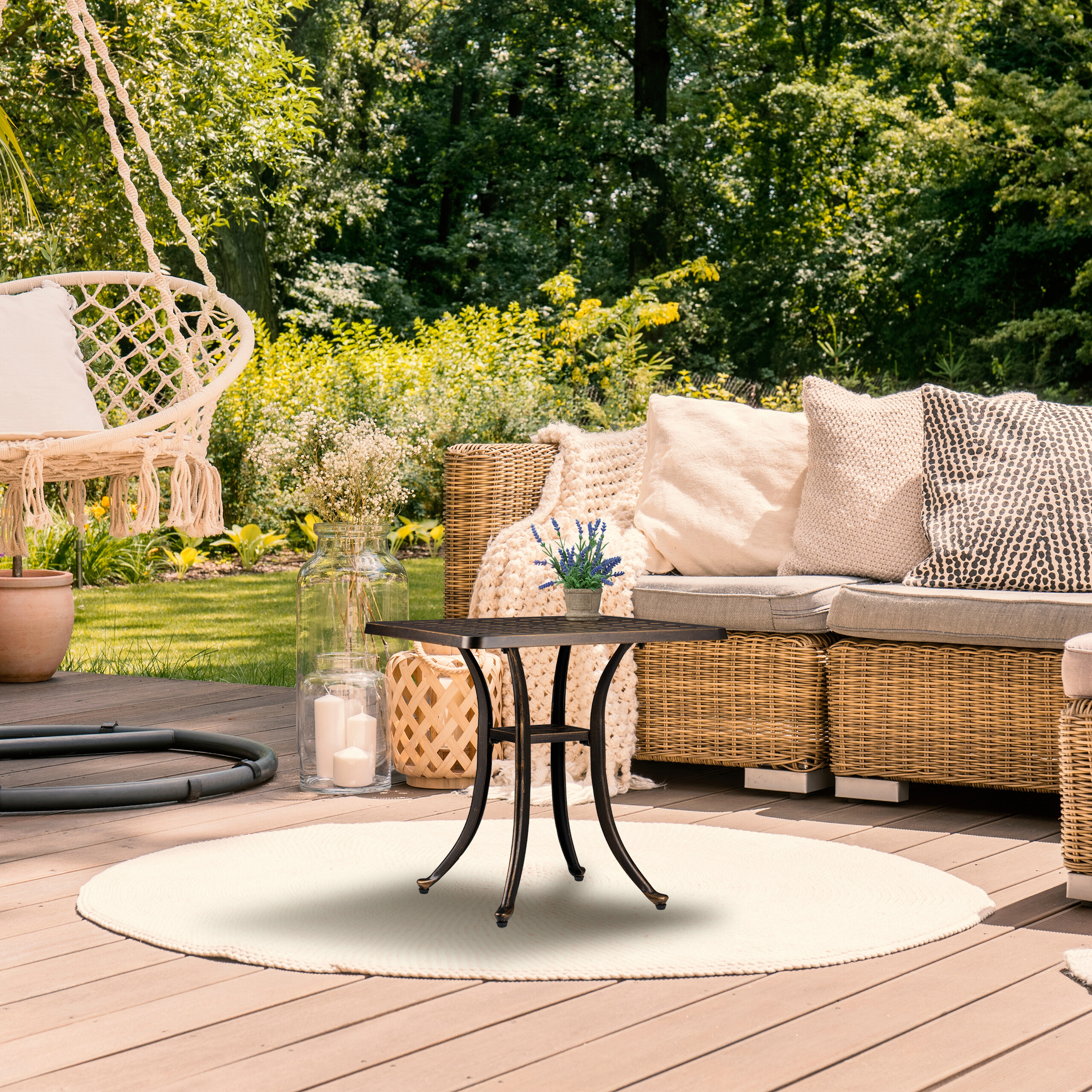 Eine Holzterrasse hat ein Weifensofa mit Kissen, einen kleinen Metalltisch, einen Hängesessel und grüne Bäume.