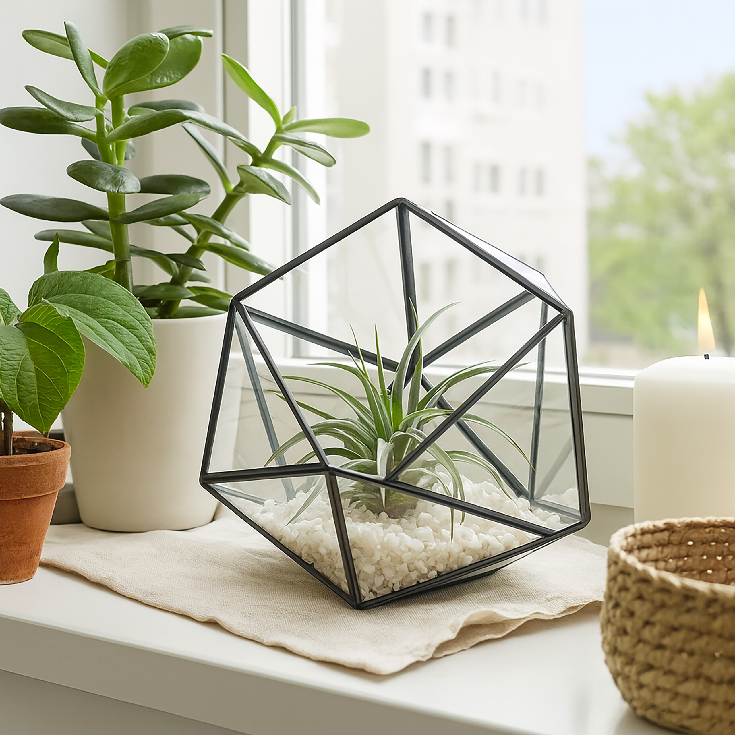 Ein Fensterbrett mit einem geometrischen Glasterrarium, Topfpflanzen, einer brennenden Kerze und einem kleinen Korb.