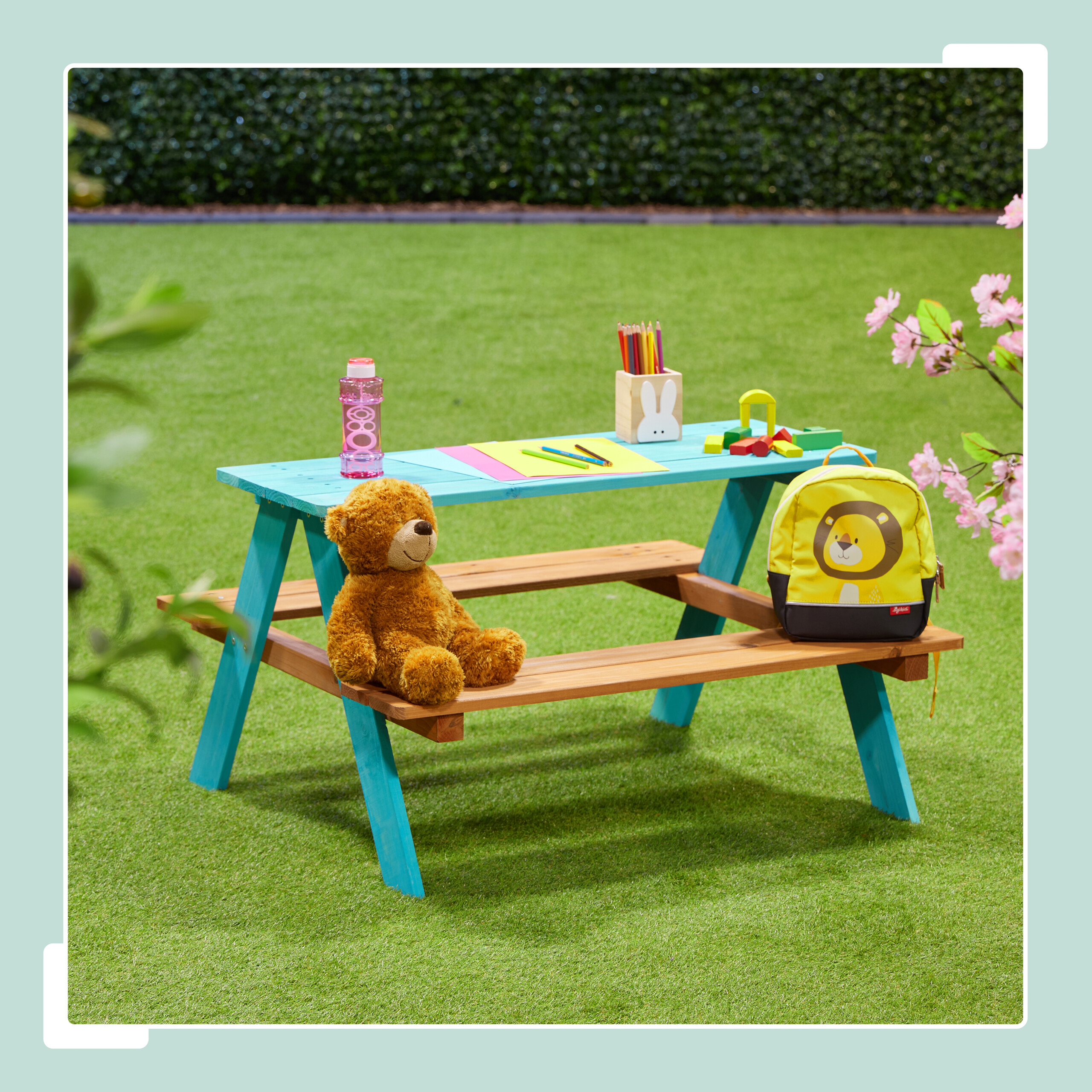Blauer Holztisch im Gras mit Teddybär, Rucksack, Wasserflasche und Stiften.