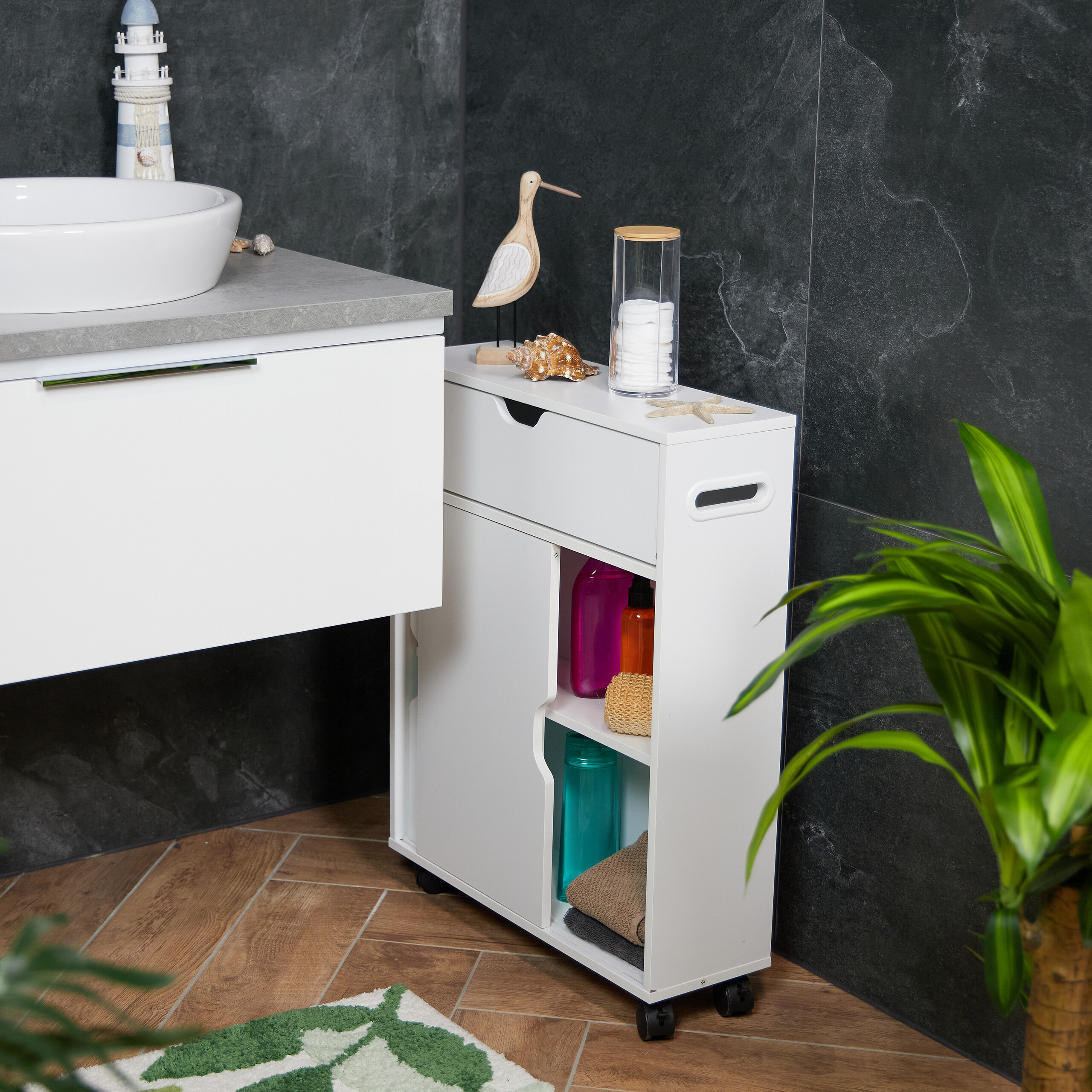 Ein weißer, schmaler Badschrank auf Rollen steht neben einem Waschbecken vor dunklen Fliesen.