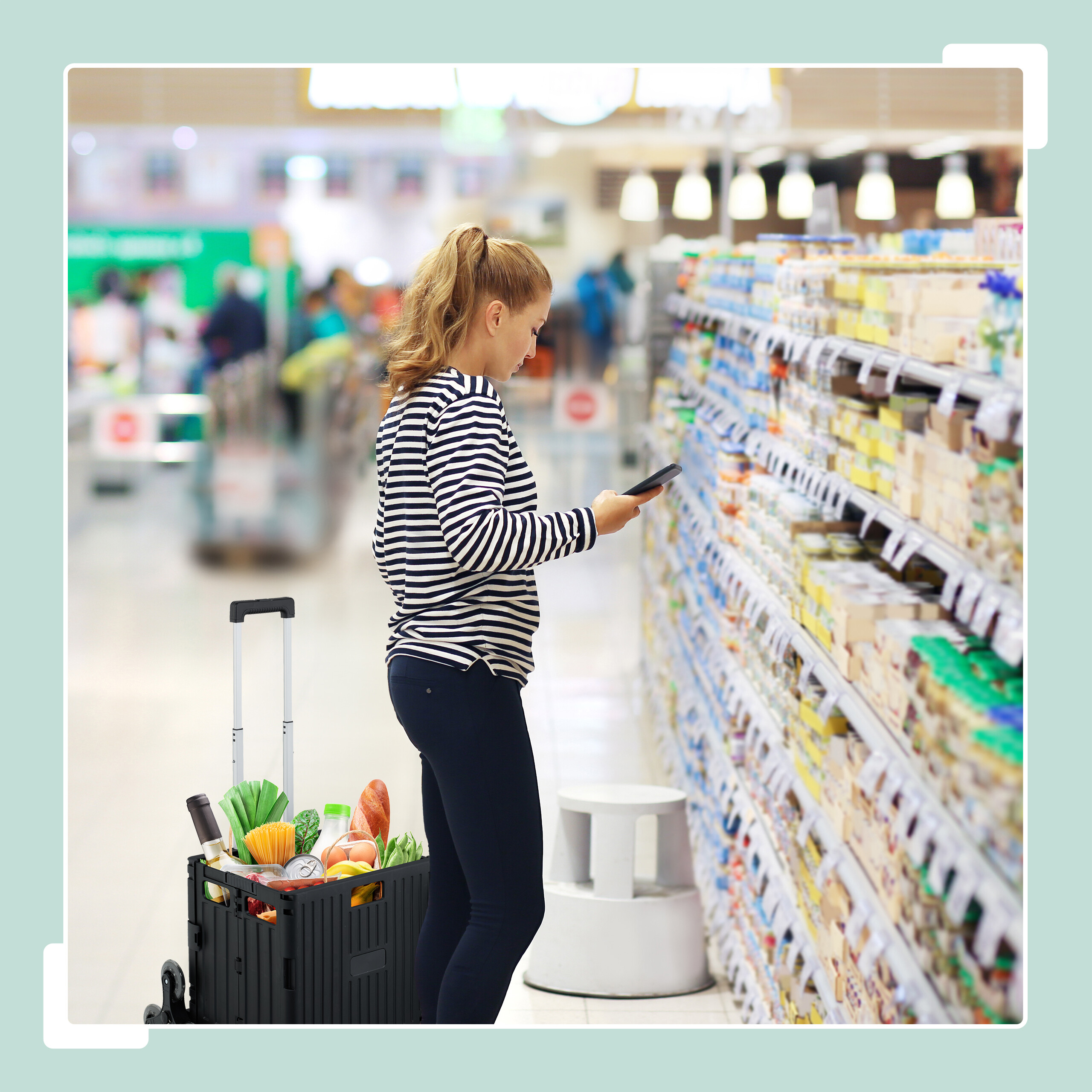 Eine Frau im gestreiften Hemd nutzt ihr Handy am Supermarktregal mit einem Einkaufswagen.