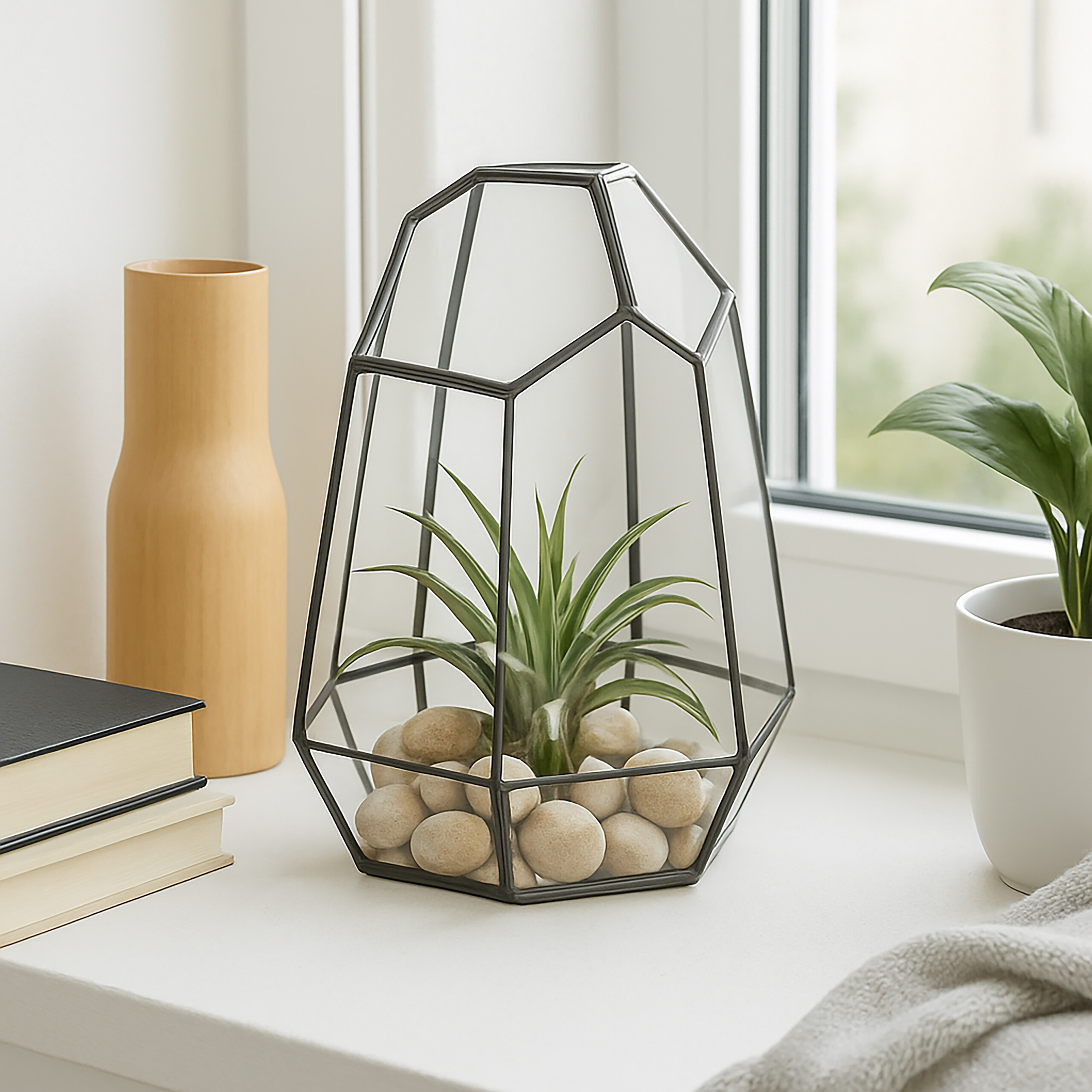 Ein geometrisches Glas-Terrarium mit einer Pflanze und Kieselsteinen auf einer Fensterbank, umgeben von Dekoration.