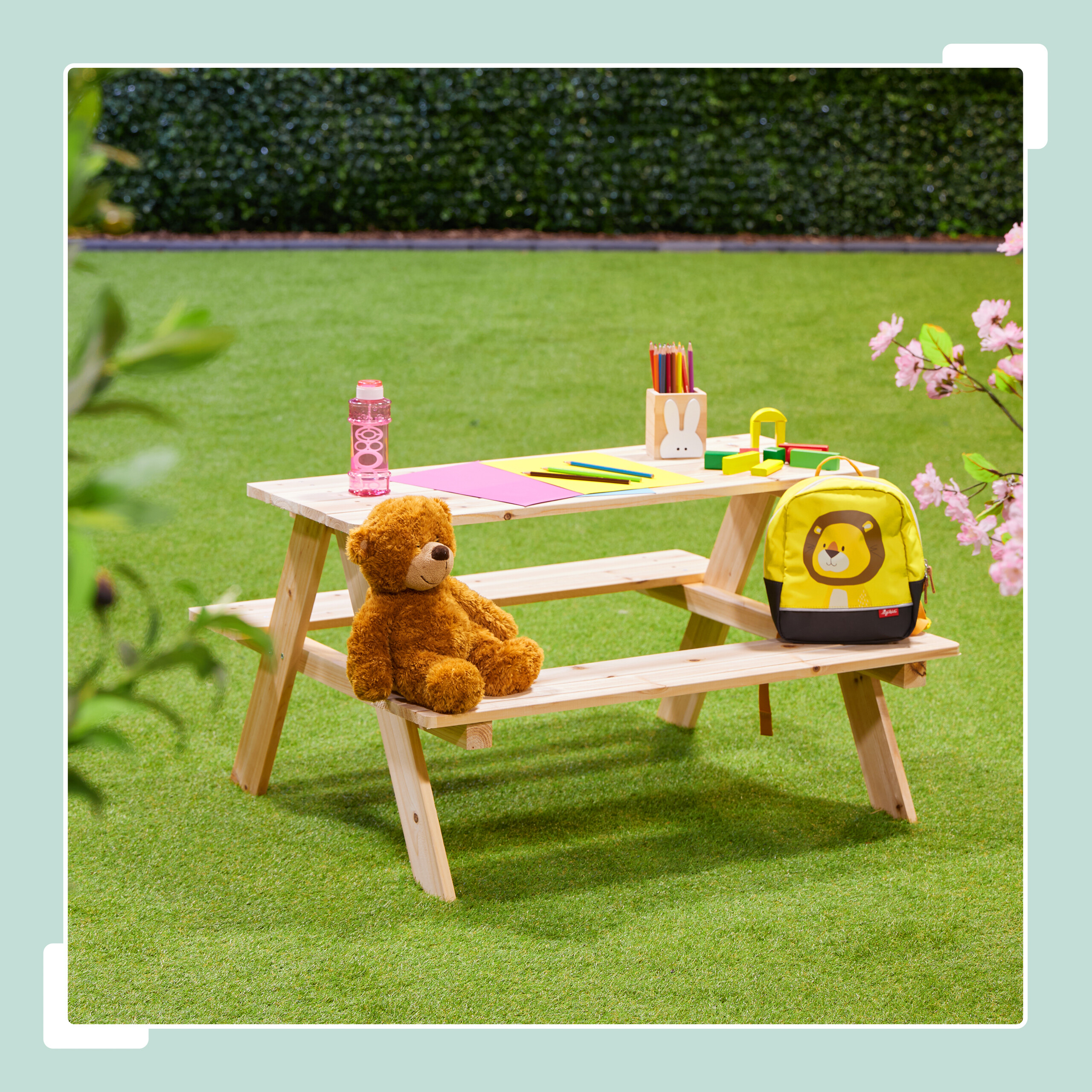 Ein Holztisch auf Rasen mit einem Teddybär, Stiftehalter, Trinkflasche und einem Löwenrucksack.