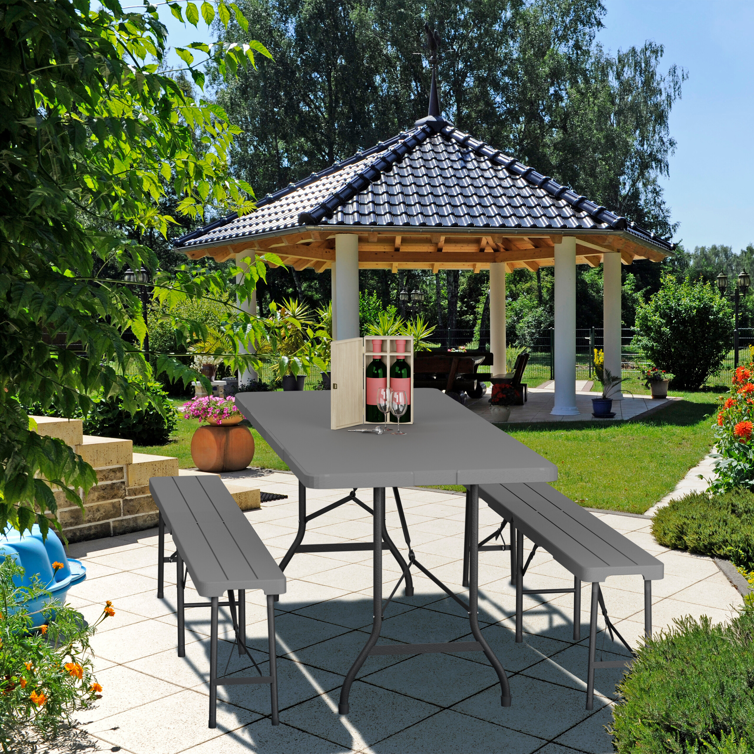 Ein grauer Klapptisch und zwei Bänke auf einer Terrasse mit einem Pavillon im Hintergrund.