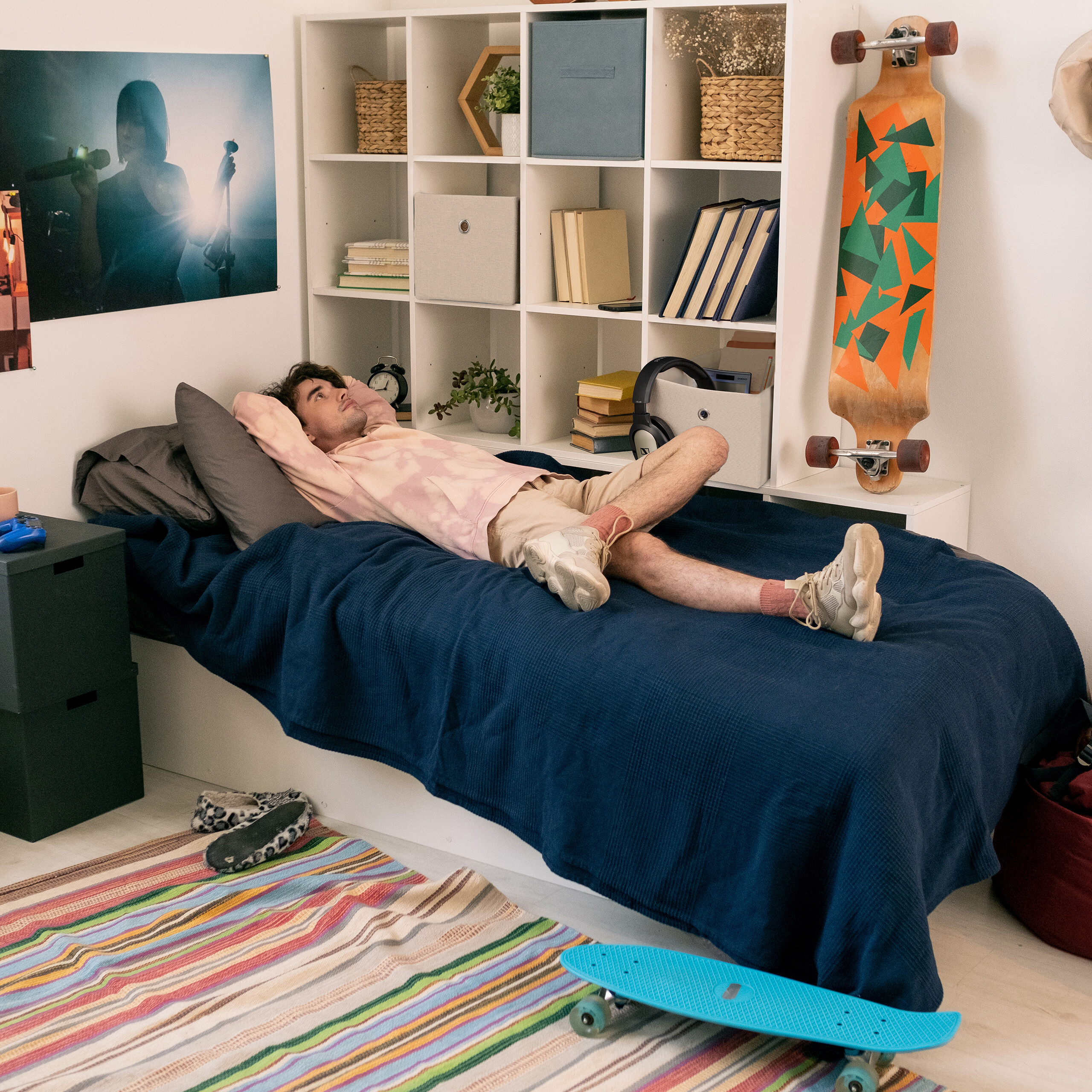Ein Mann im rosa Hoodie entspannt in einem Zimmer mit weißem Regal und Longboard an der Wand auf einem blauen Bett.
