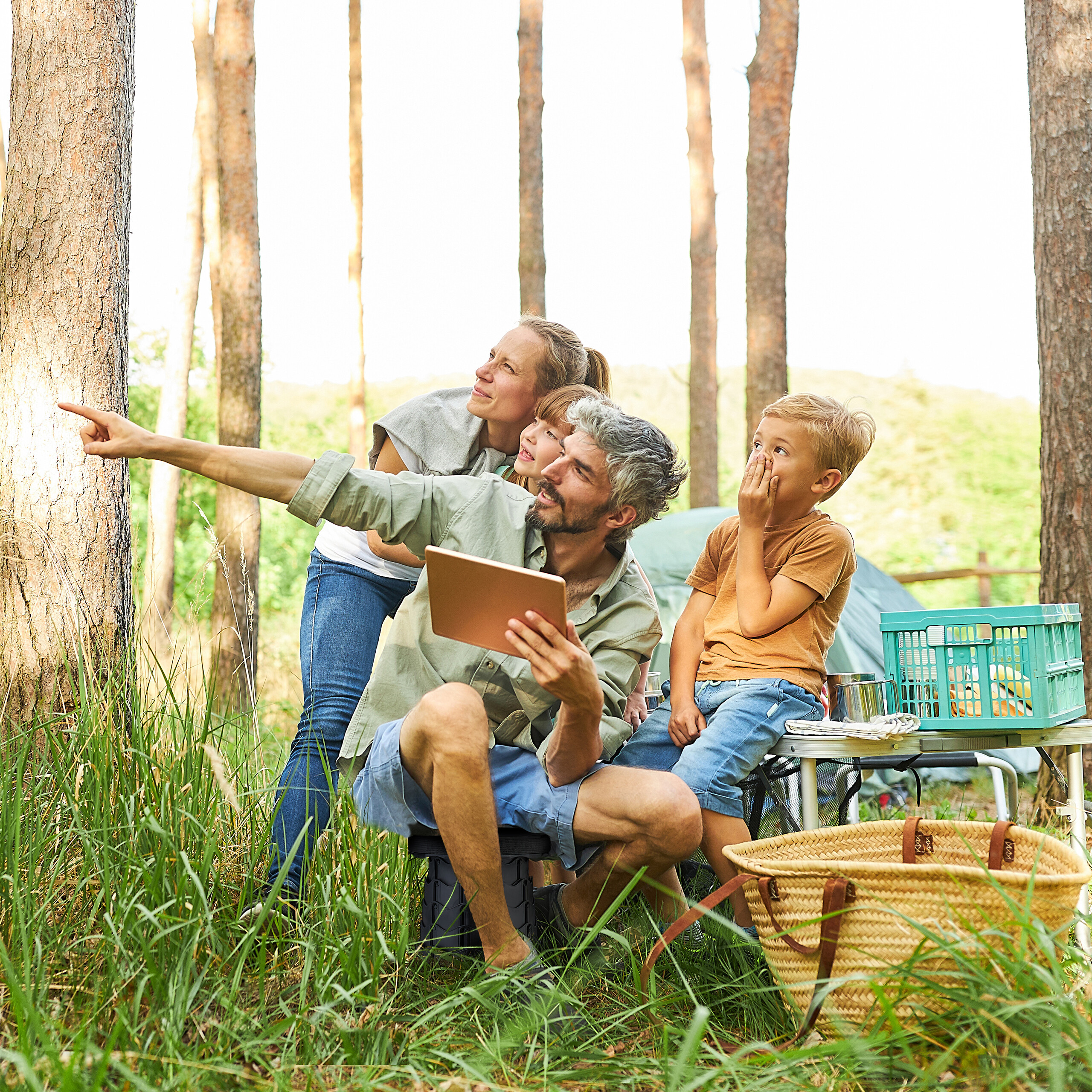 Eine Familie beim Camping im Wald, der Vater hält ein Tablet und zeigt auf etwas, die Mutter und Kinder schauen zu.