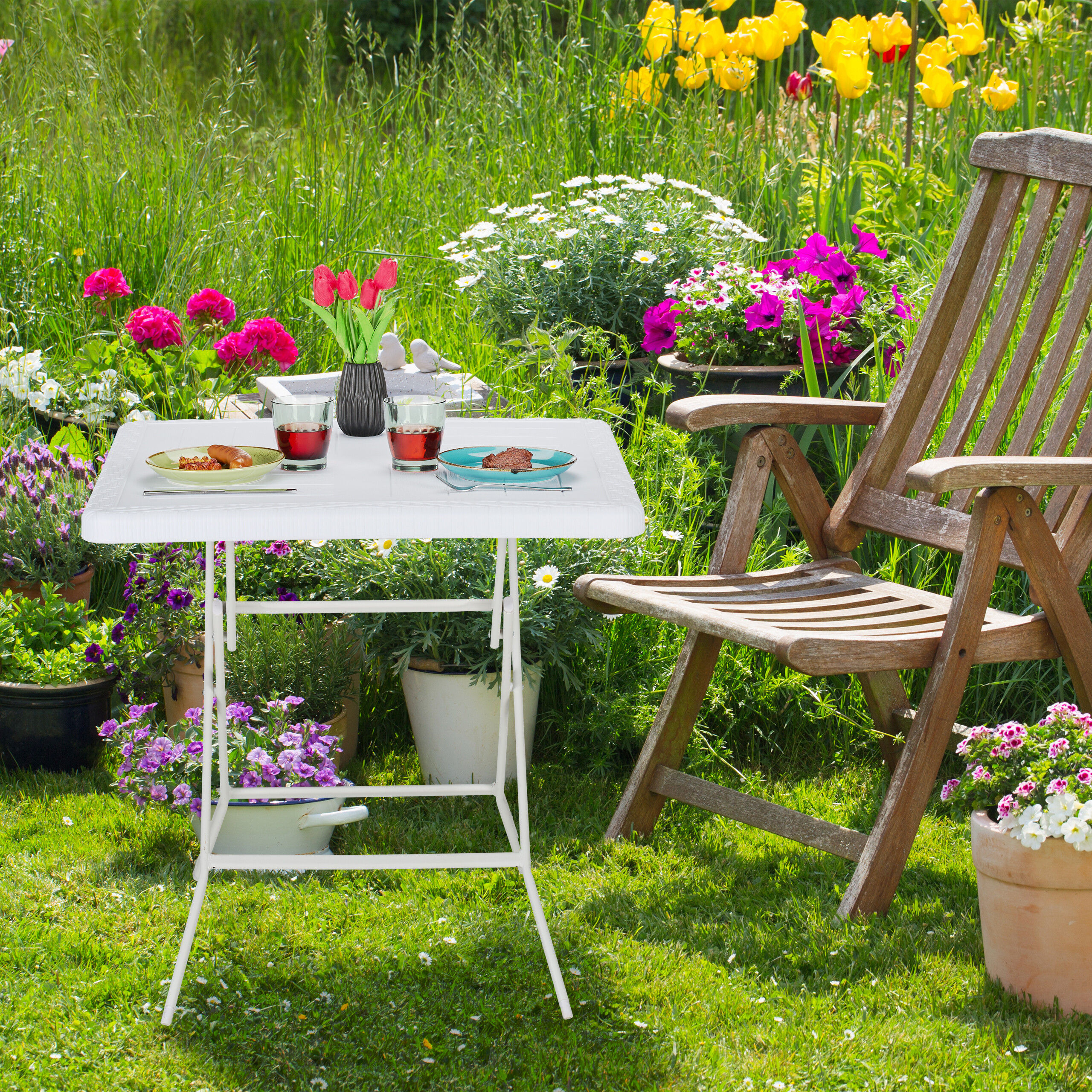 Ein kleiner weißer Tisch mit Essen, Getränken und Blumen steht auf einer grünen Wiese neben einem Holzstuhl.