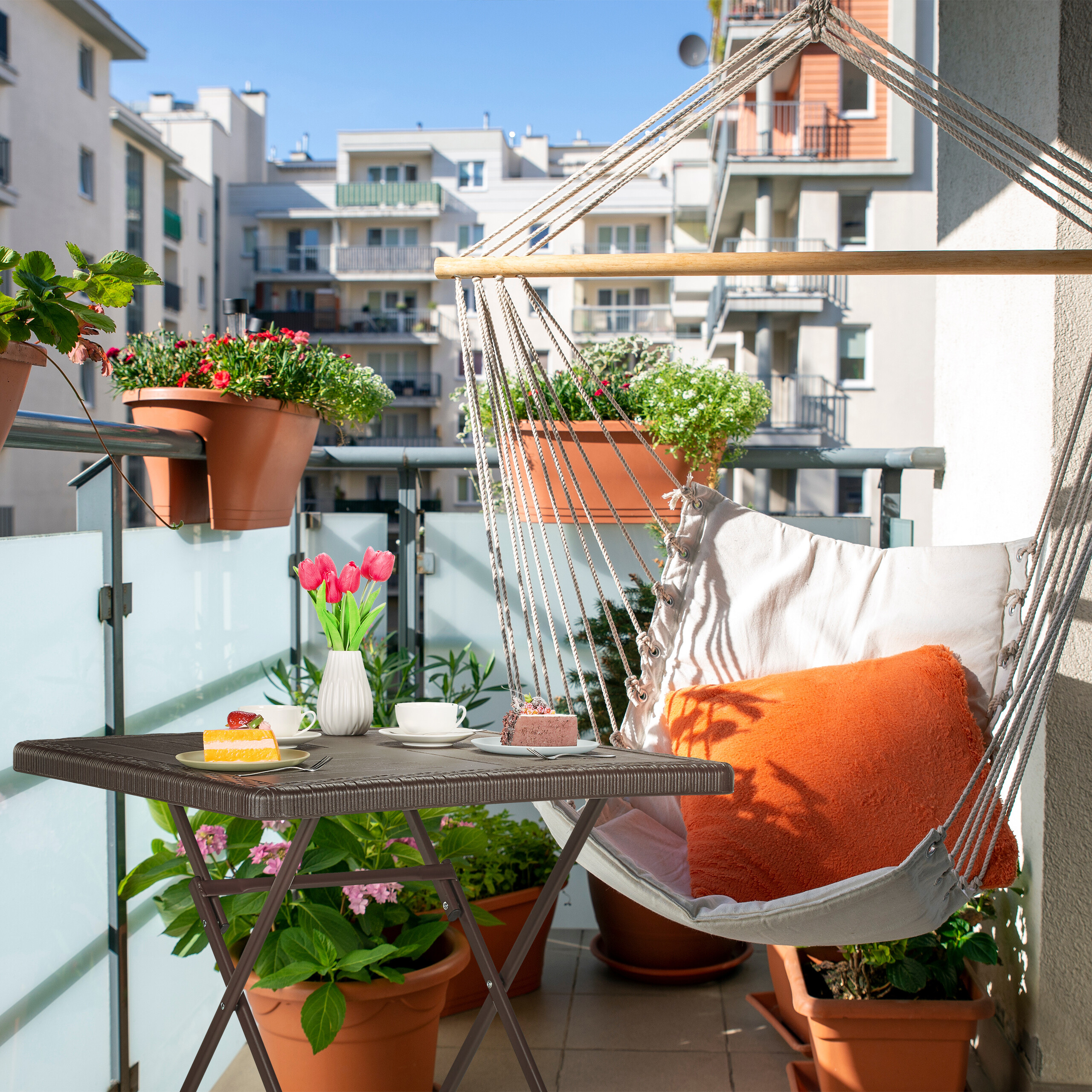 Ein weißer Hängesessel mit Kissen auf einem sonnigen Balkon mit Tisch, Blumen und Blick auf Gebäude.