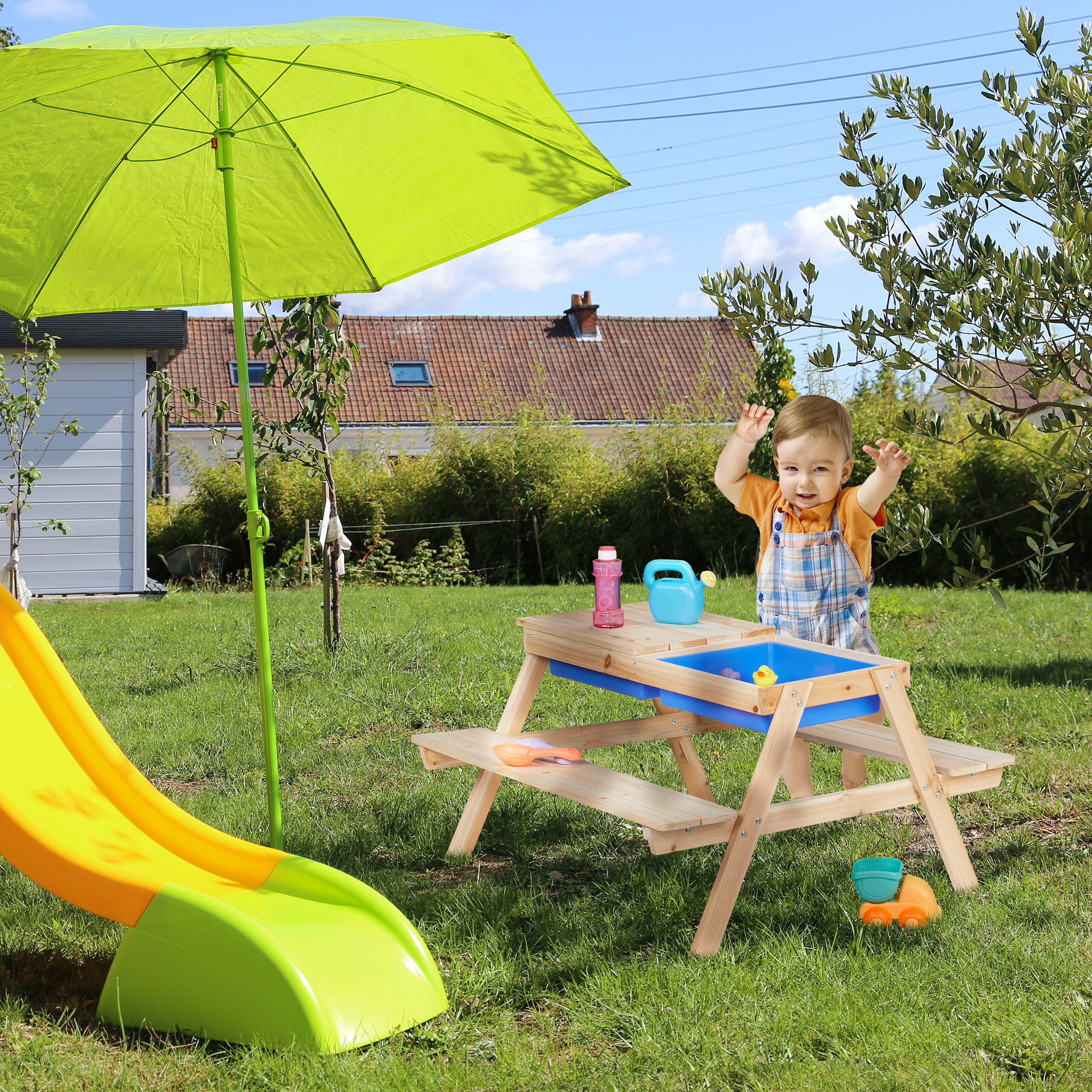 Ein Kleinkind spielt an einem Holztisch mit Spielzeug im sonnigen Garten mit grünem Schirm und Rutsche.