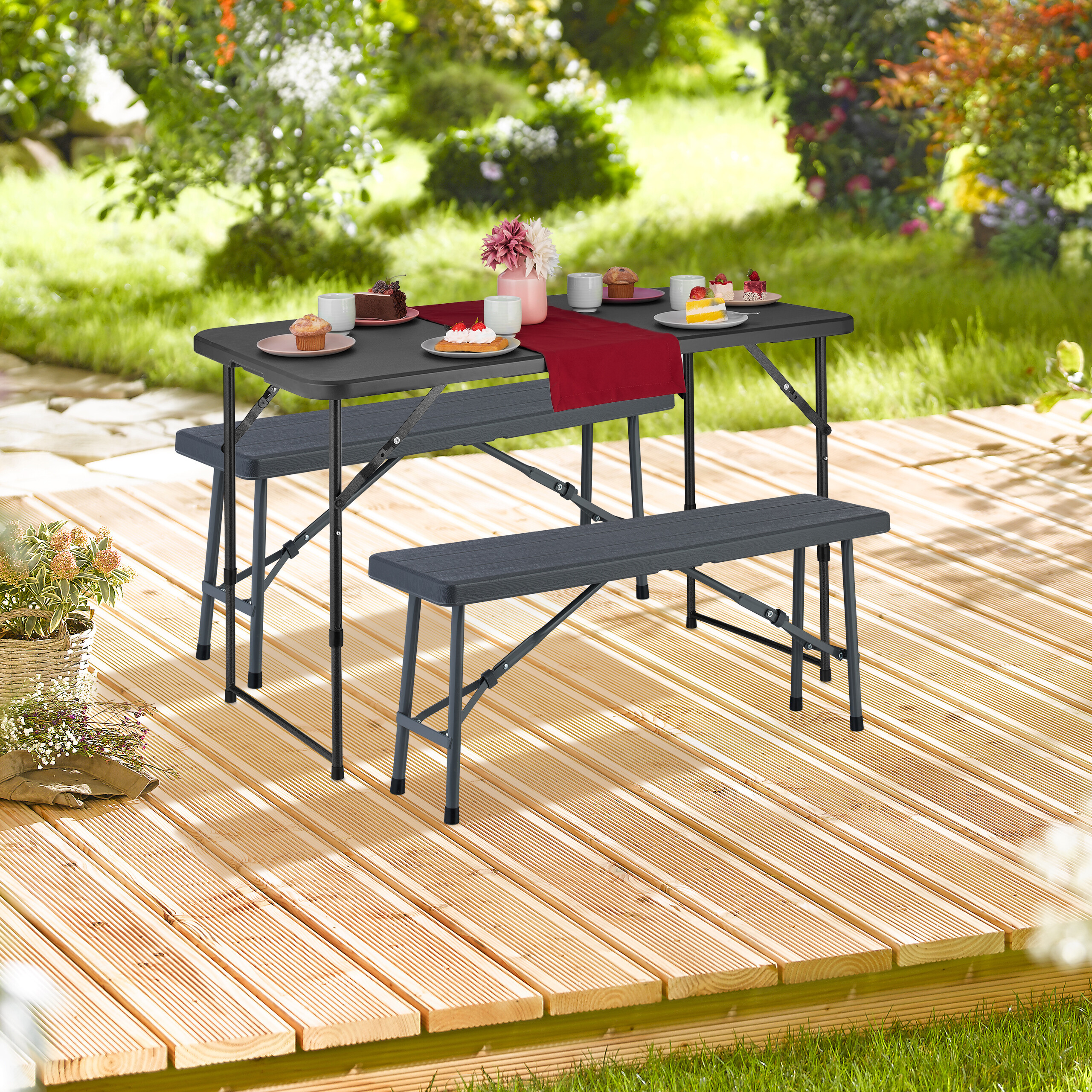 Ein schwarzer Klapptisch mit Bänken auf Holzdeck im Garten, gedeckt mit Kuchen, Tassen und Blumen.