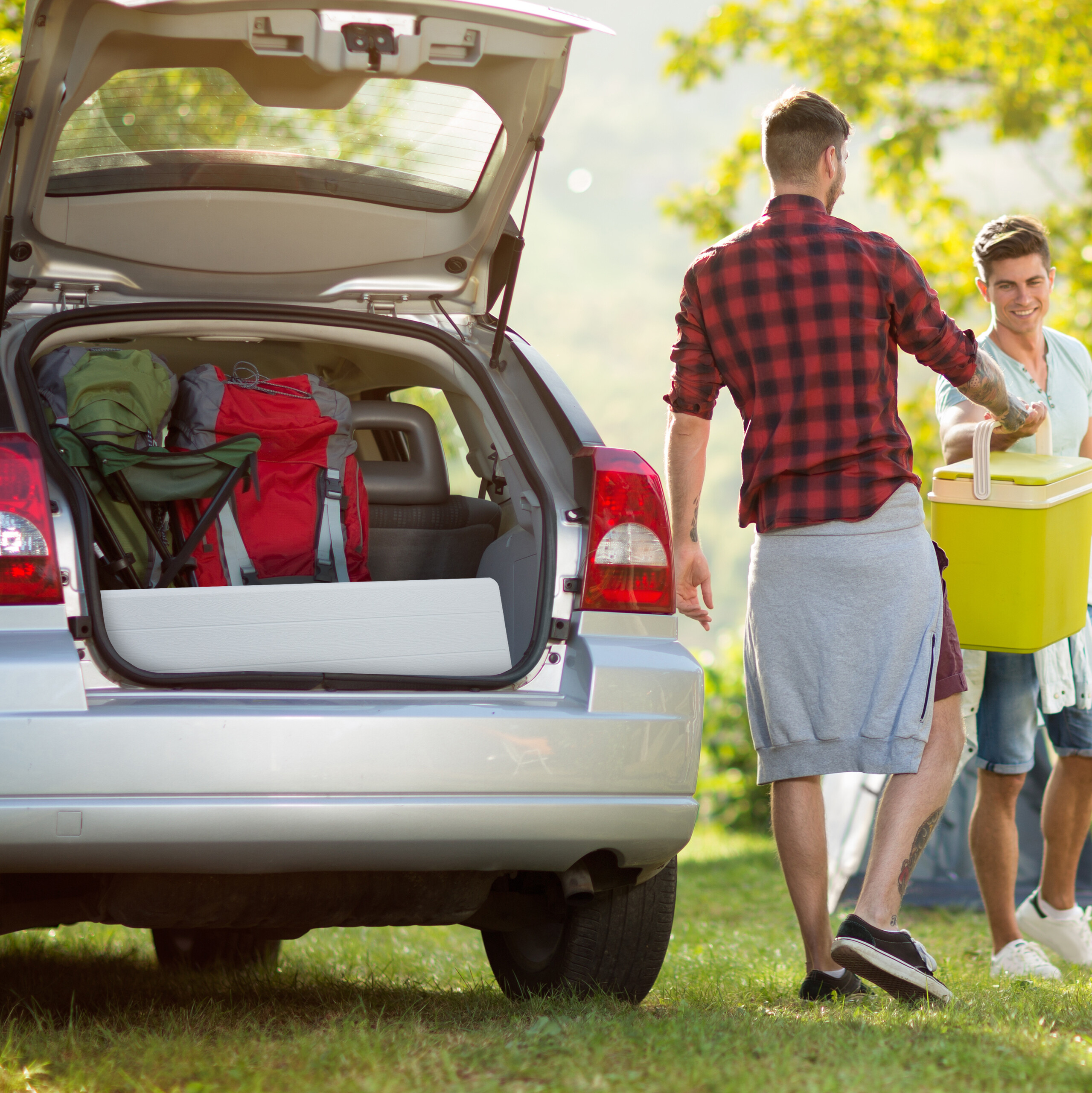 Zwei junge Männer packen Rucksäcke und eine Kühlbox aus dem Kofferraum eines Autos in der Natur aus.