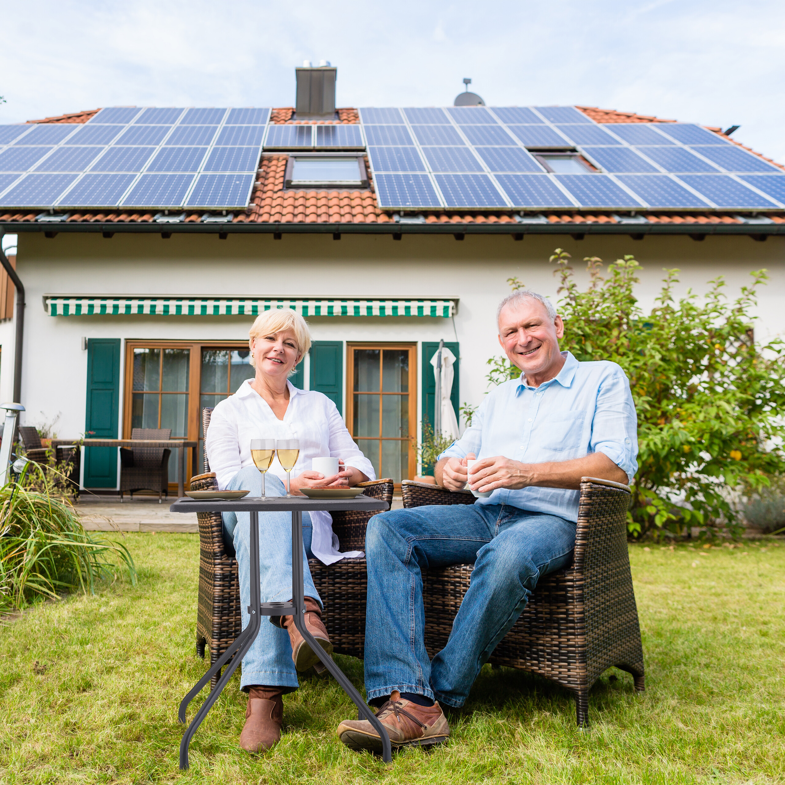 Älteres Paar sitzt entspannt im Garten vor einem Haus mit Solaranlagen auf dem Dach.